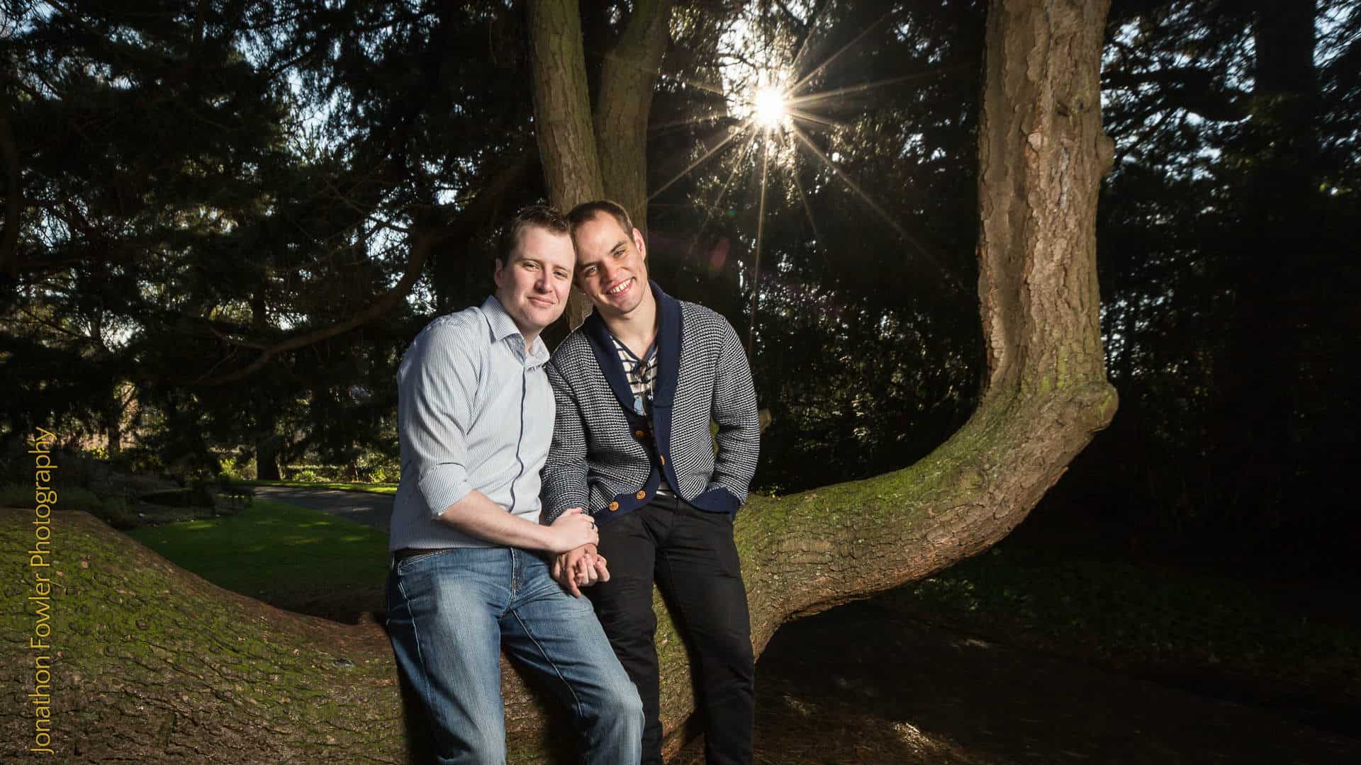 Two men smiling and holding hands, leaning against a tree in a park with sunlight filtering through the branches.
