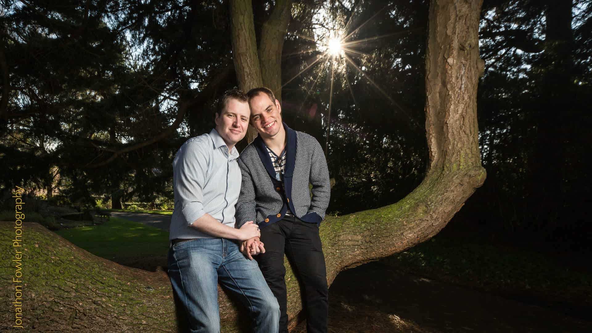 Two men smiling and holding hands, leaning against a tree in a park with sunlight filtering through the branches.