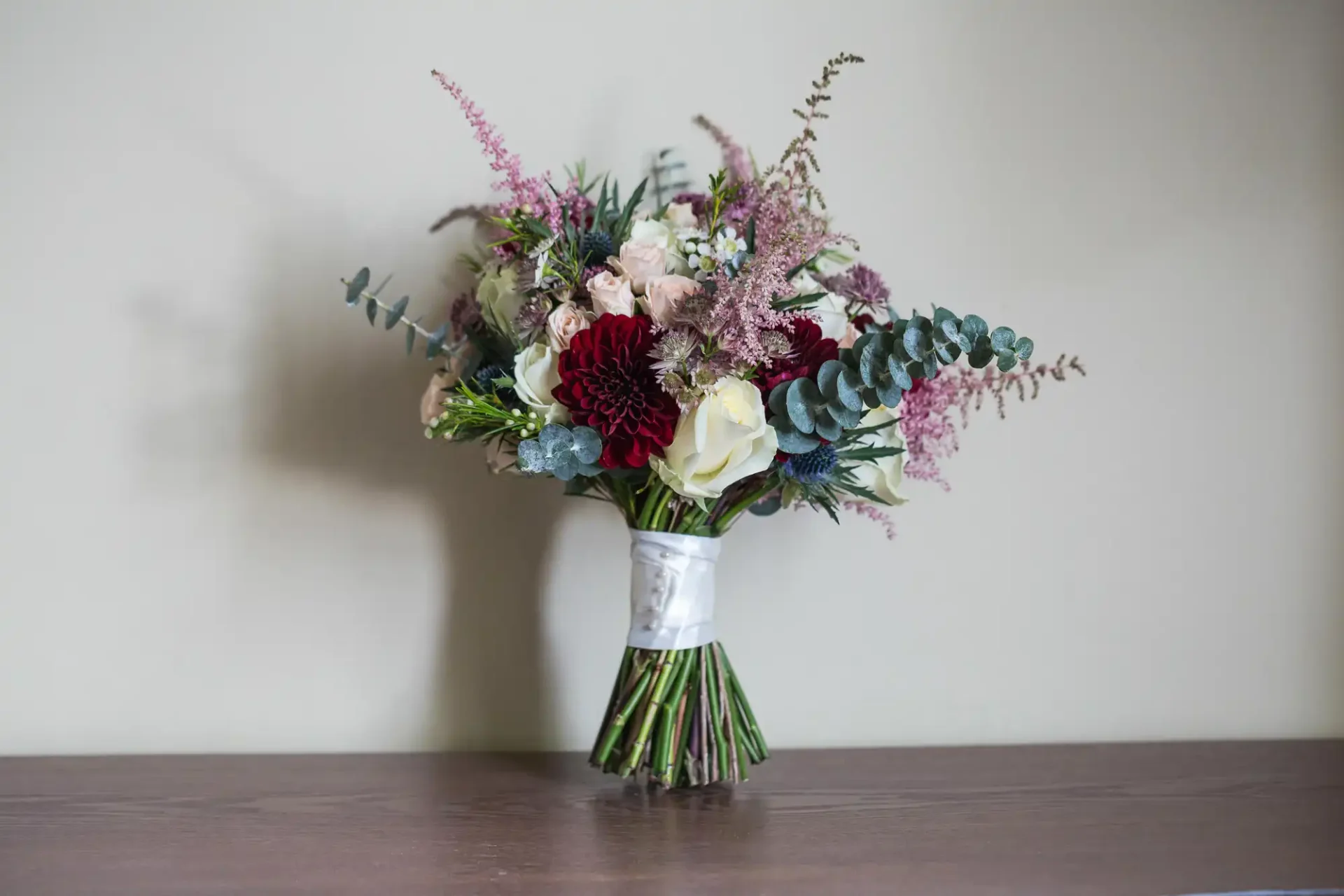 A bouquet of mixed flowers including pink, white, and deep red blooms with green foliage, wrapped in white paper, resting on a wooden surface.