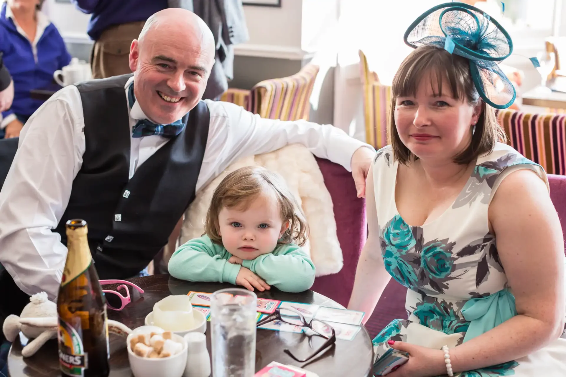 A family of three (man, woman, and toddler) smiling at a dining table decorated with drinks and snacks during a formal event.