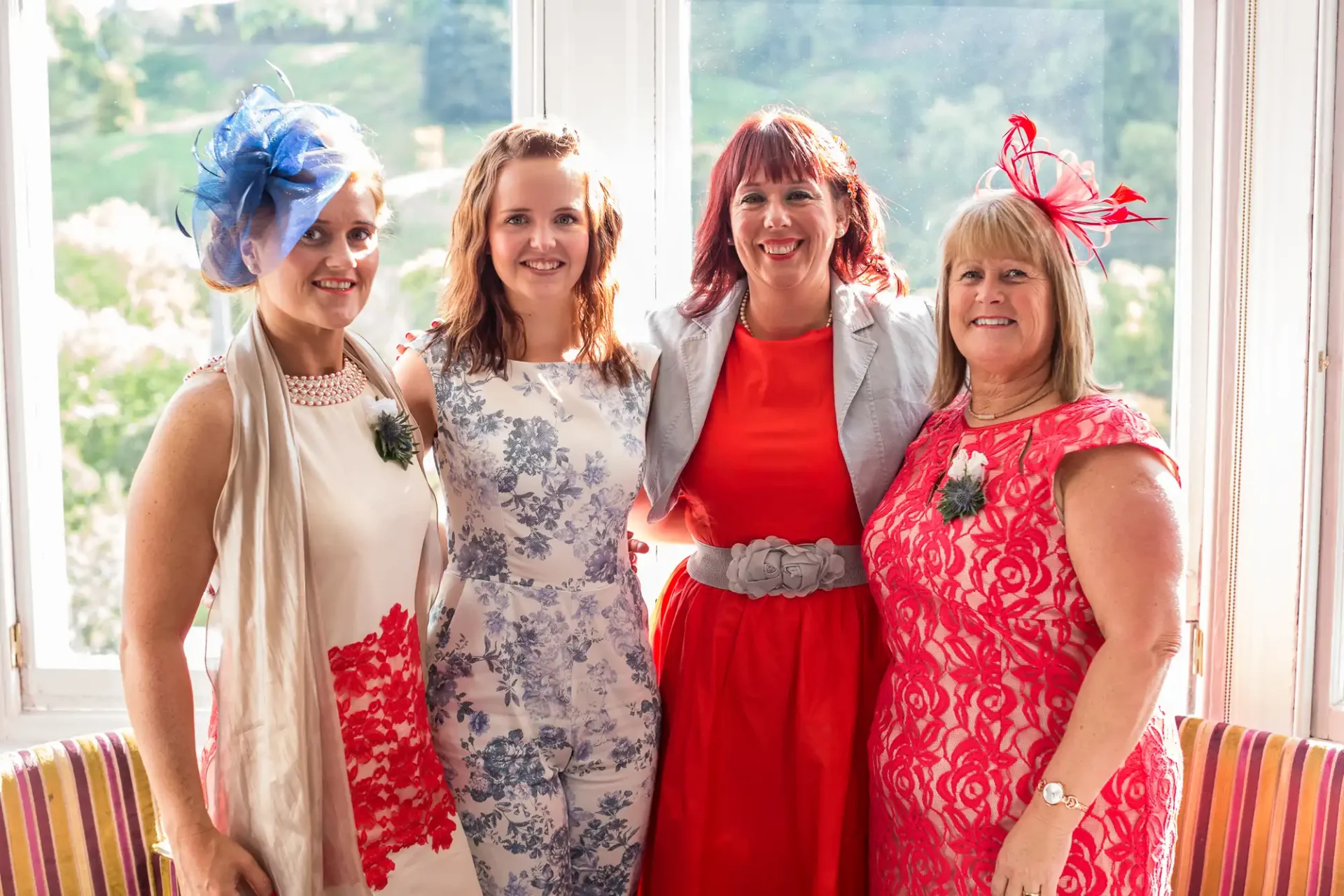 Four women wearing formal dresses and colorful hats posing together in a sunlit room.
