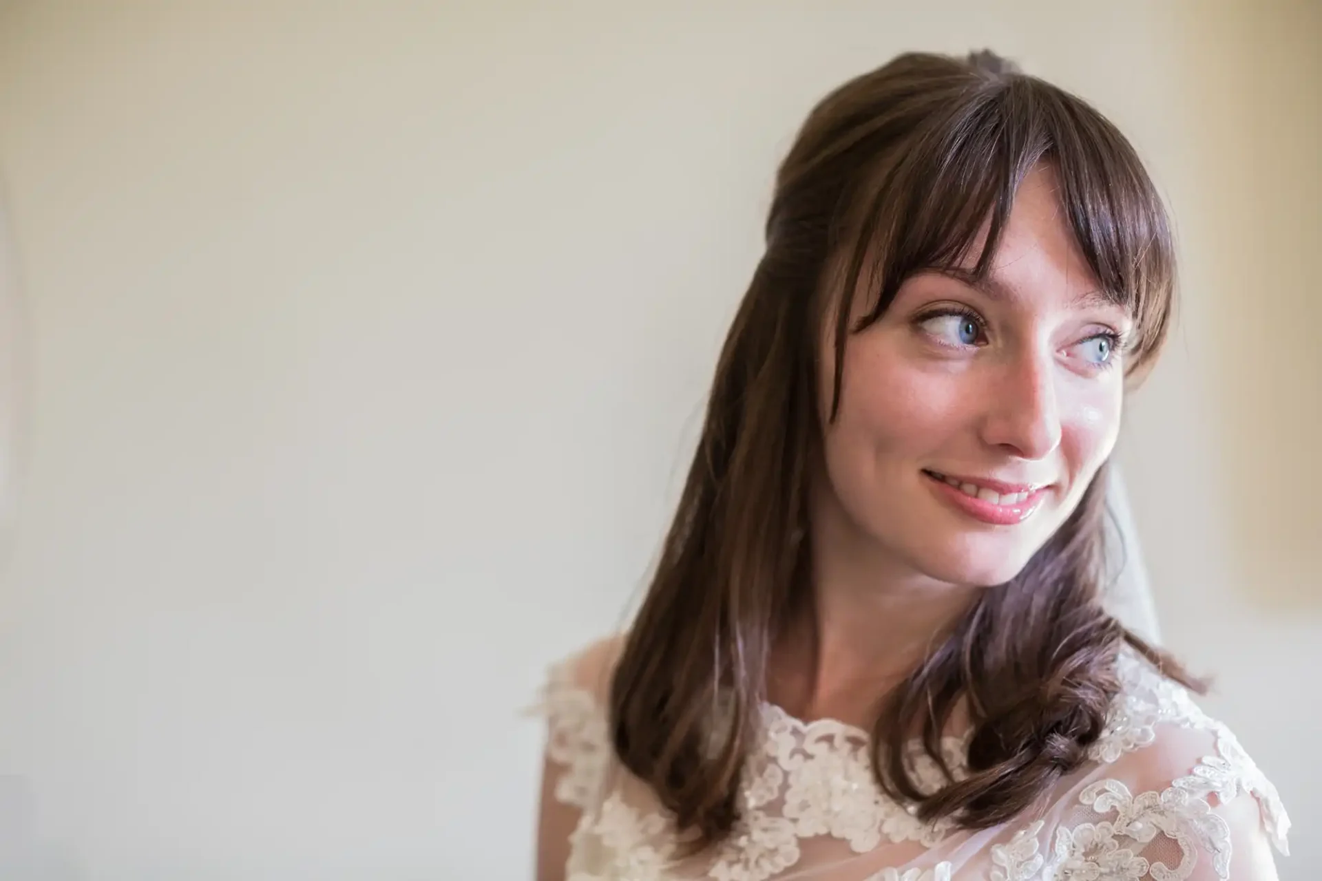 A woman with brown hair and blue eyes, wearing a white lace dress, smiles gently while looking to the side.