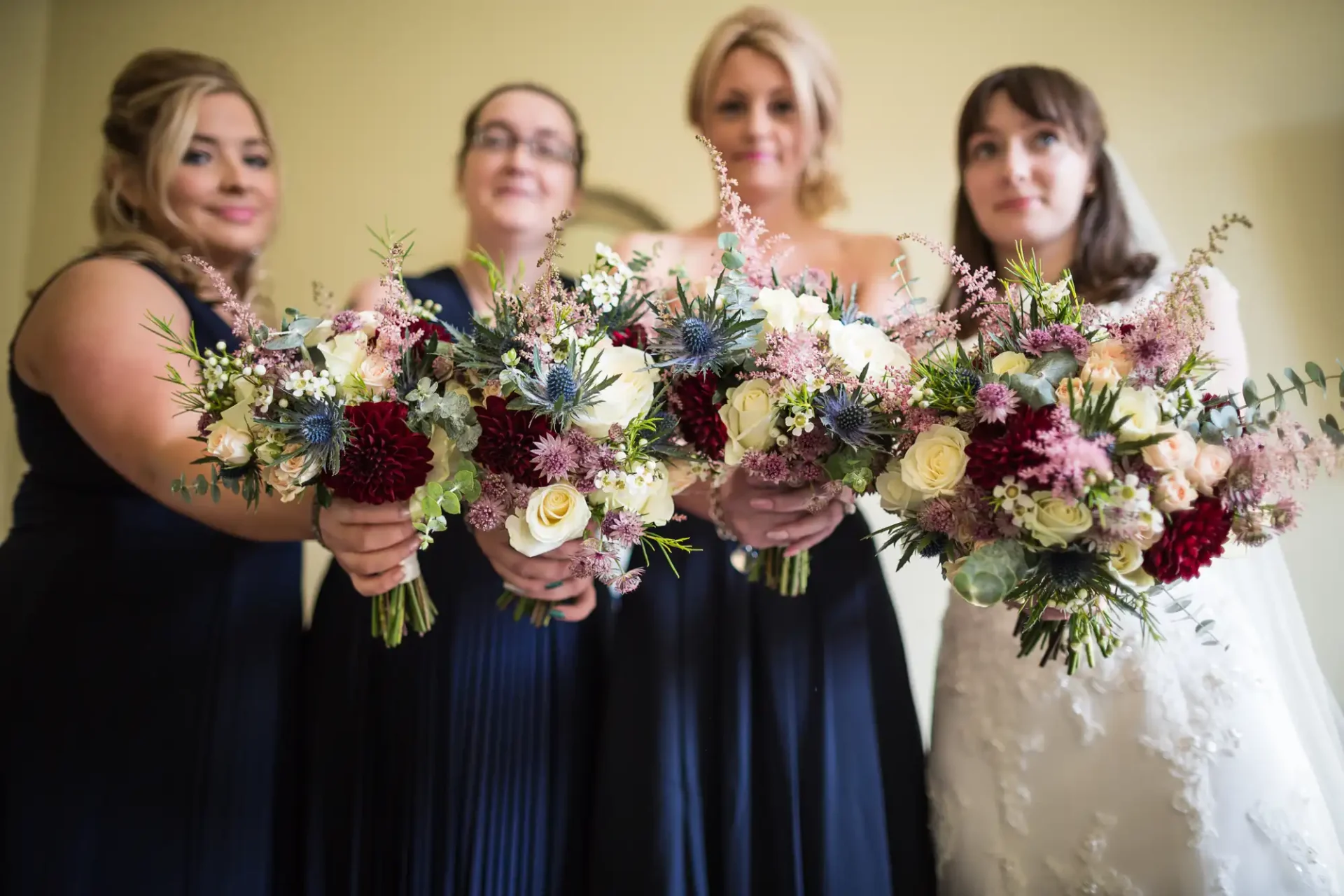 Four women in formal attire hold intricate flower bouquets towards the camera in a room with a yellow background.