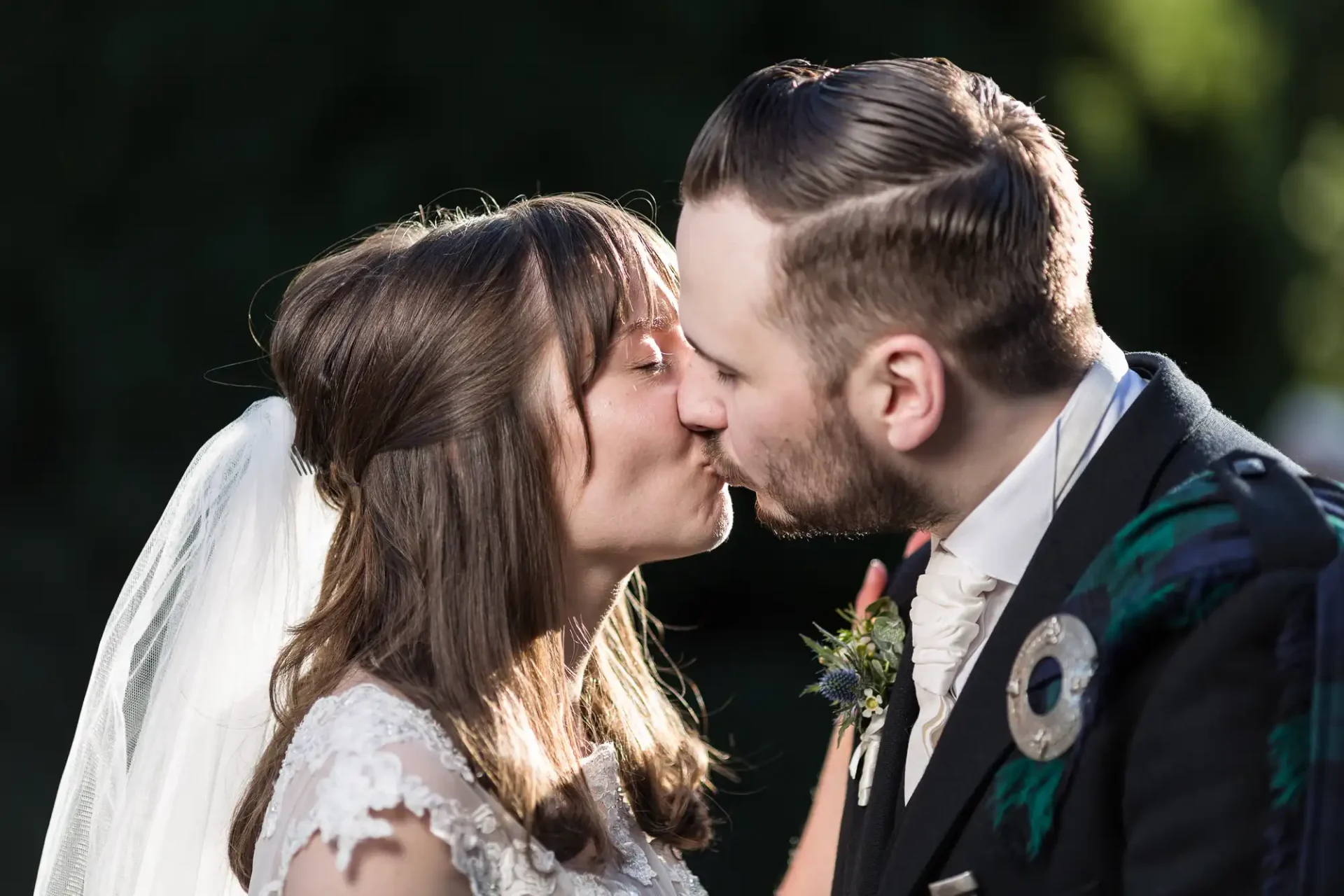 A bride and groom kissing, both dressed in traditional wedding attire, with a sunlit natural backdrop.