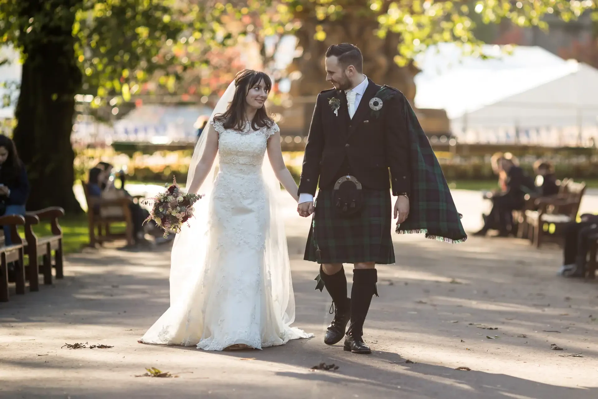 Bride and groom walking hand in hand in a park, the bride in a white dress and the groom in a kilt, with sunlight filtering through trees.