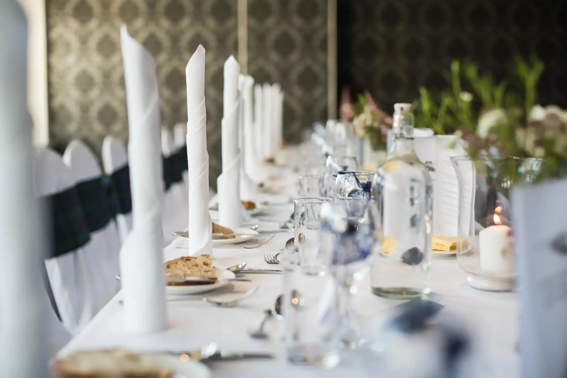 Elegant dining table set with white linen, tall napkins, silverware, and clear water bottles, in a room with a patterned wallpaper.