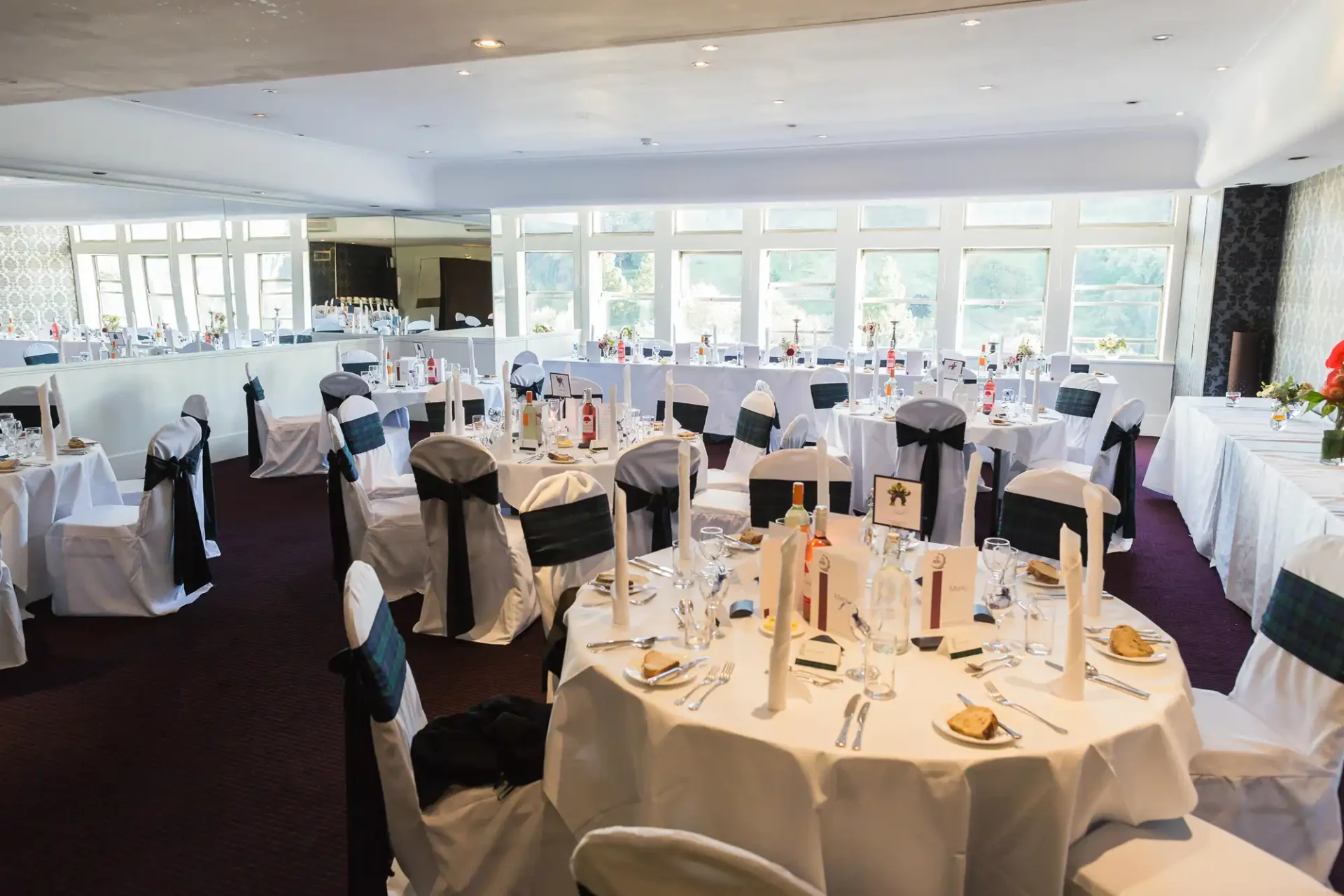 Elegant dining room set for an event with white-covered tables, black chairs with white sashes, and large windows overlooking greenery.