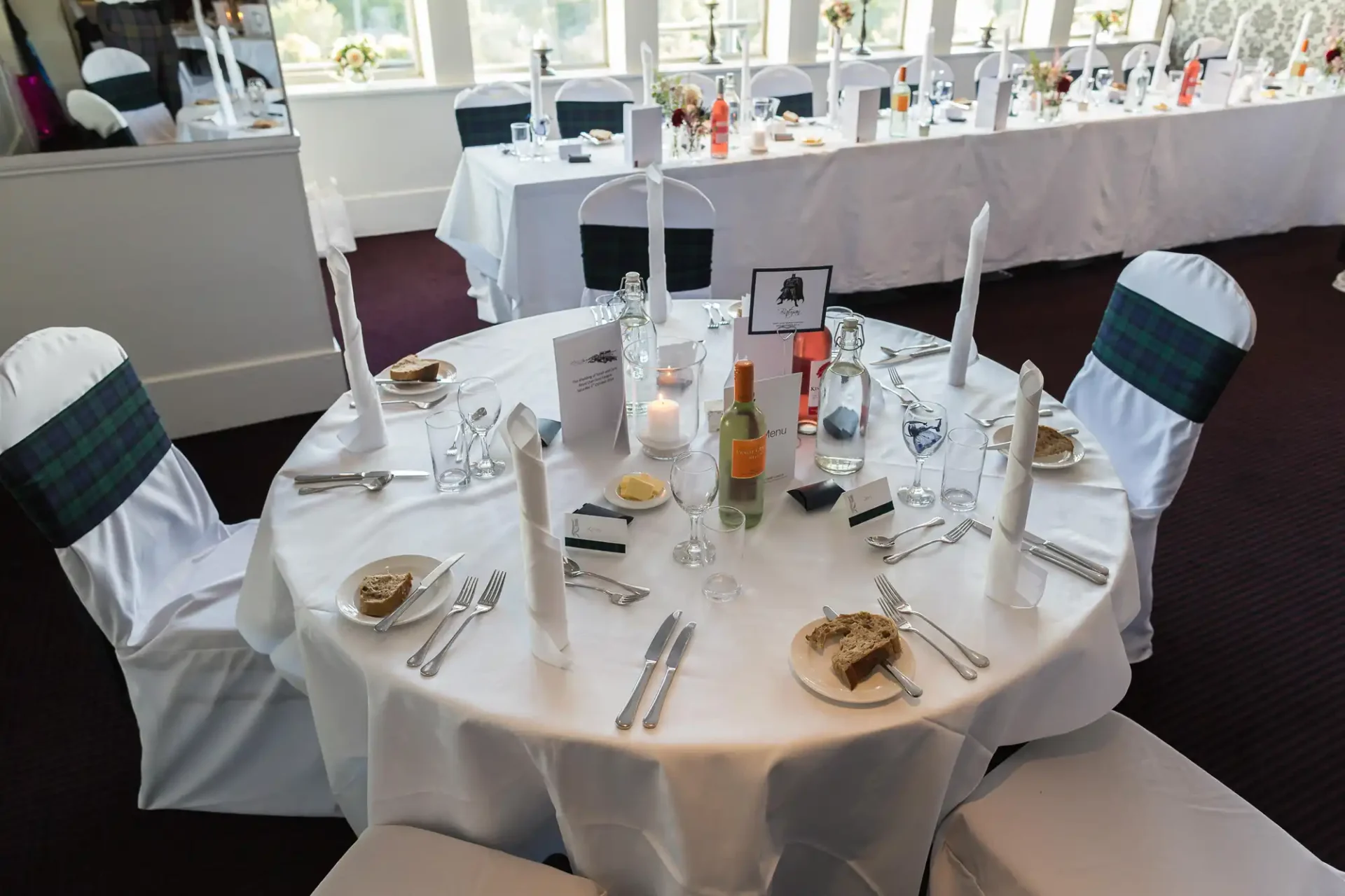 Elegantly set wedding reception table with white linens, wine glasses, name cards, and menu cards in a brightly lit room.