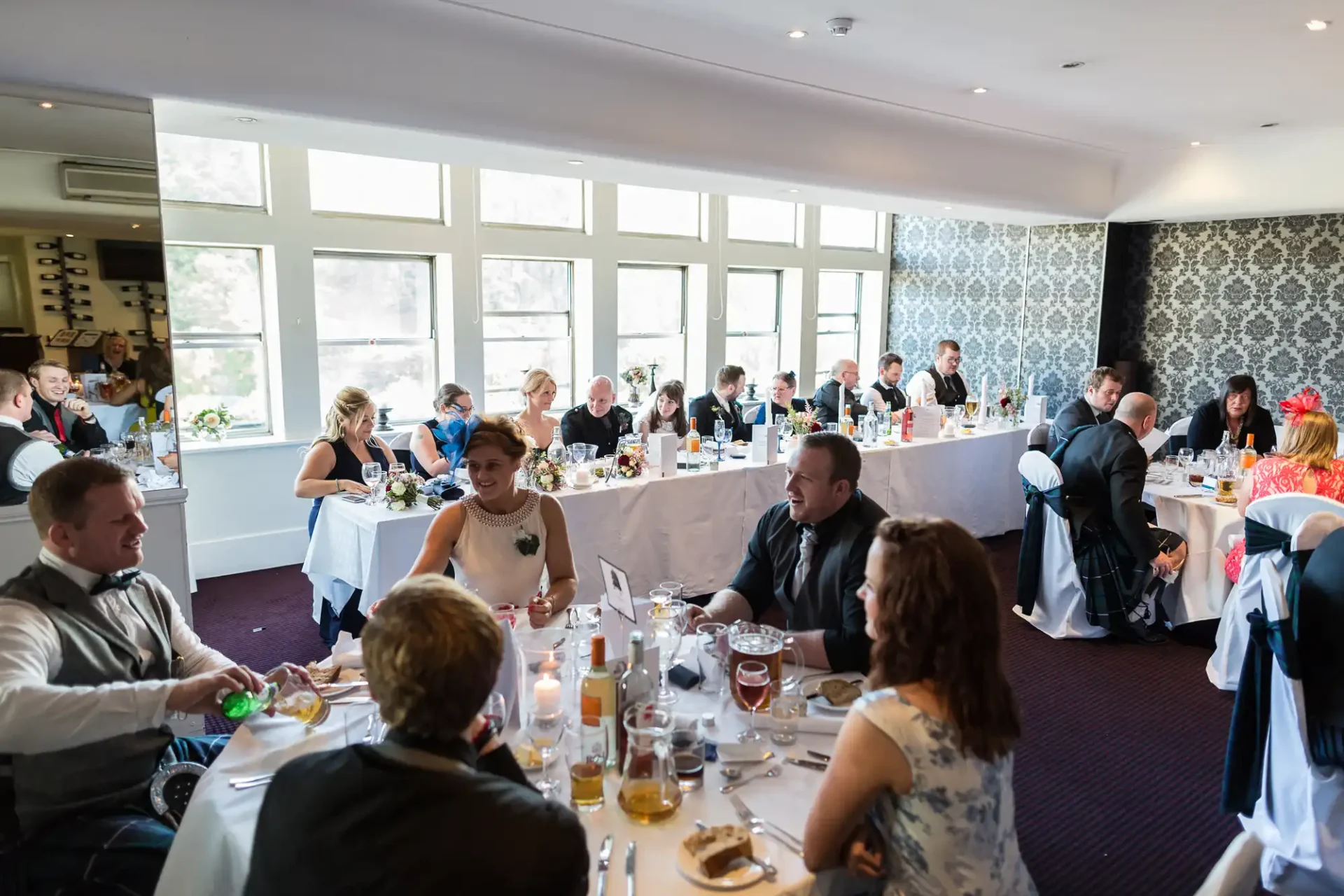 Guests seated at tables during a wedding reception, chatting and dining in an elegantly decorated room with large windows.