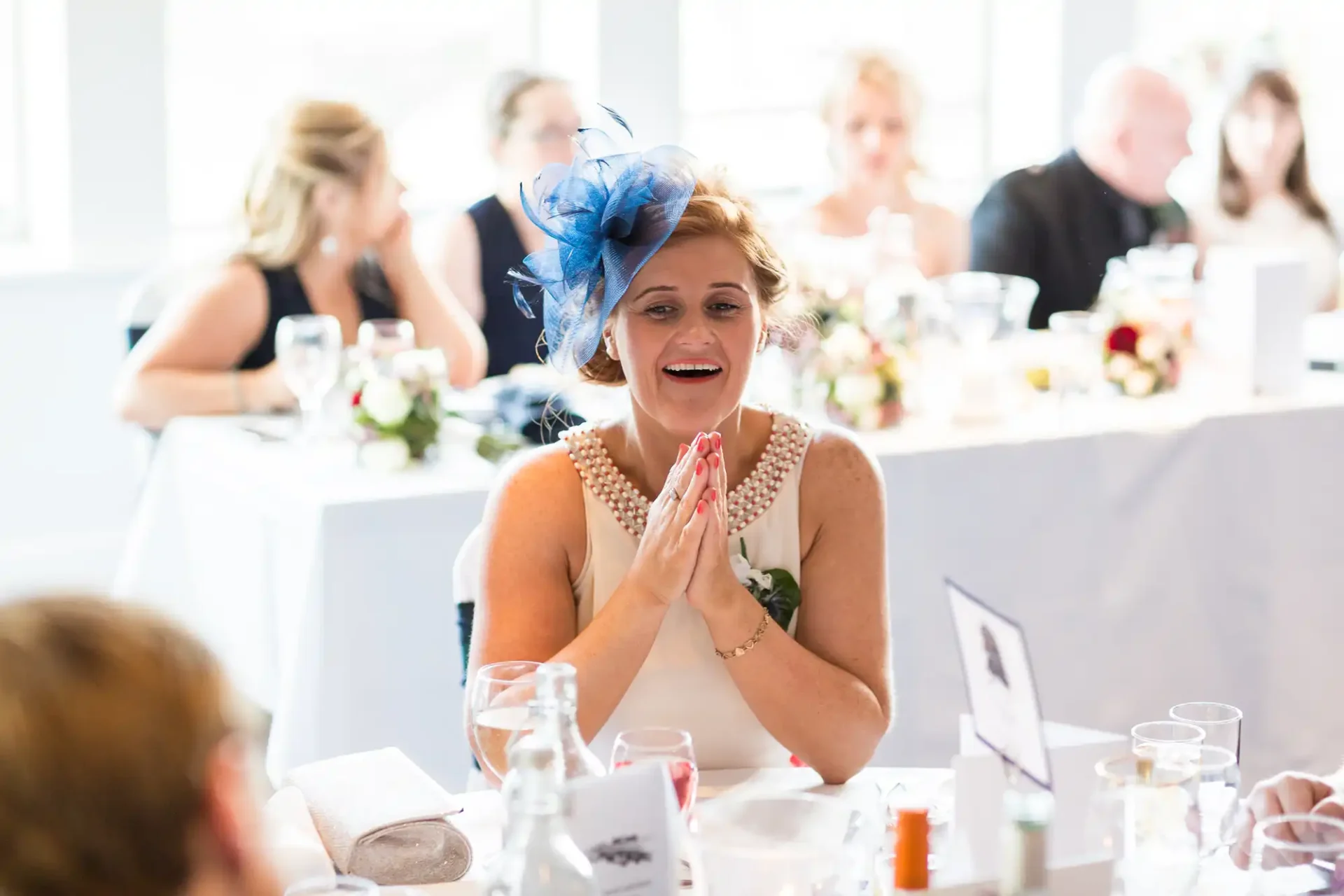 A woman in a festive hat and pearl necklace looks surprised at a wedding reception, clapping her hands together with guests in background.