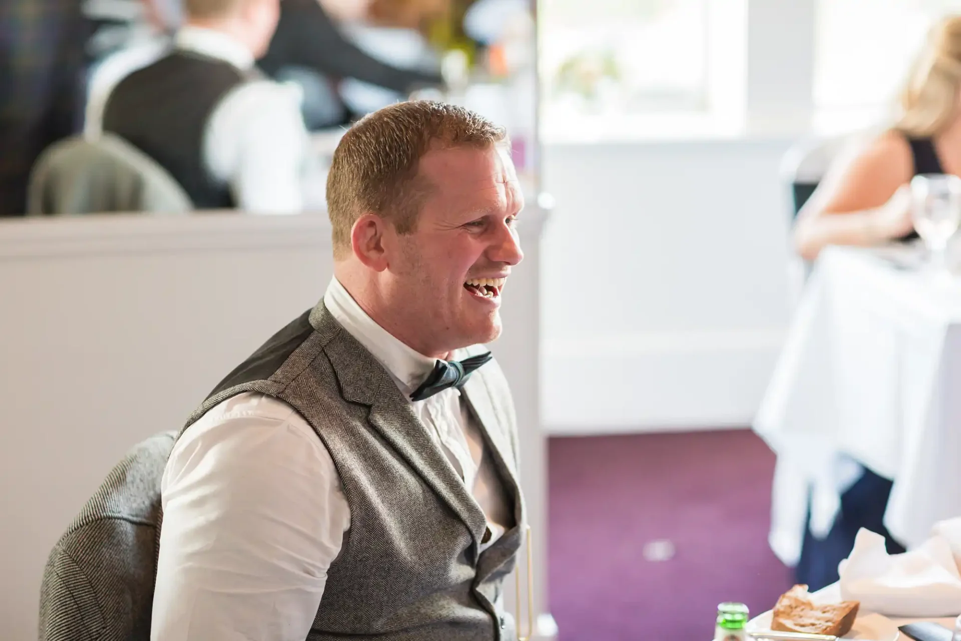 Man in a gray vest and bow tie laughing at a formal event, with blurred guests in the background.