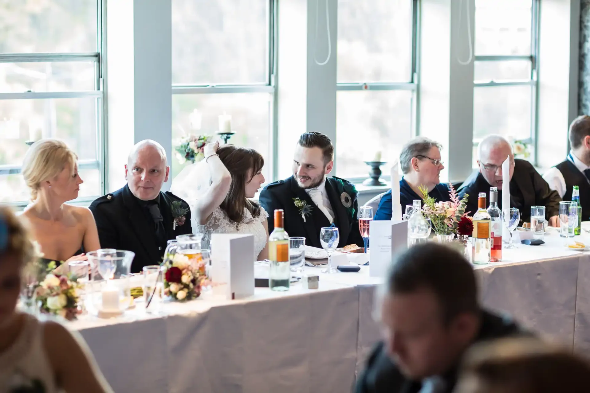 A wedding reception scene inside a well-lit venue with large windows, showing guests seated at a table decorated with flowers, engaging in conversation.
