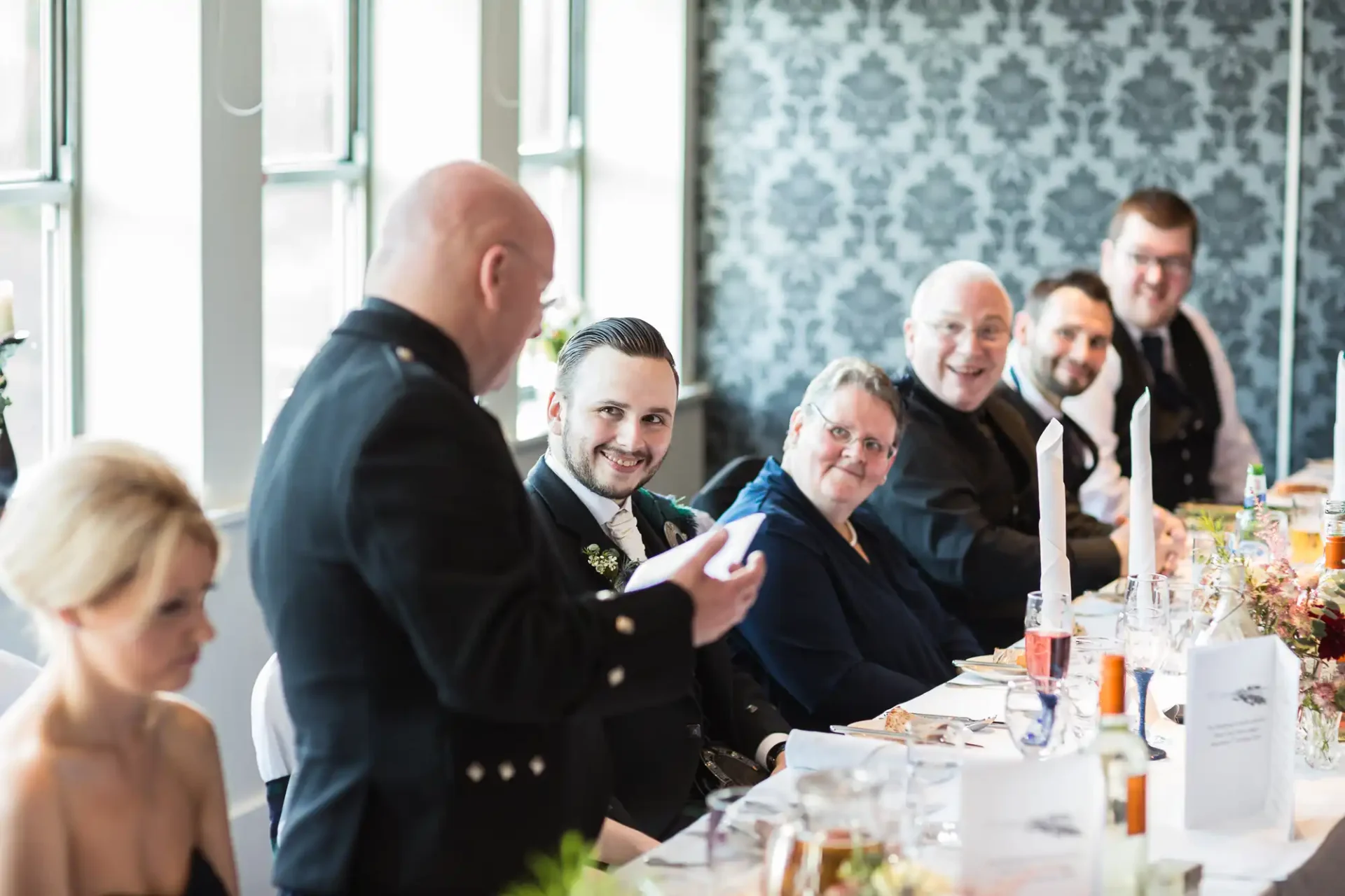 A man giving a speech at a wedding reception, holding a microphone, with guests seated at the table smiling and listening.