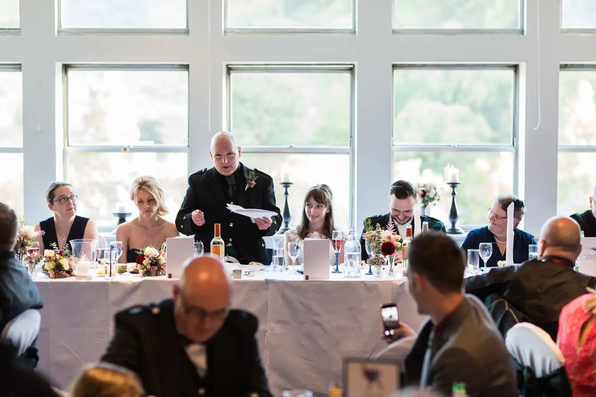 A man stands giving a speech at a wedding table surrounded by seated guests in a room with large windows.