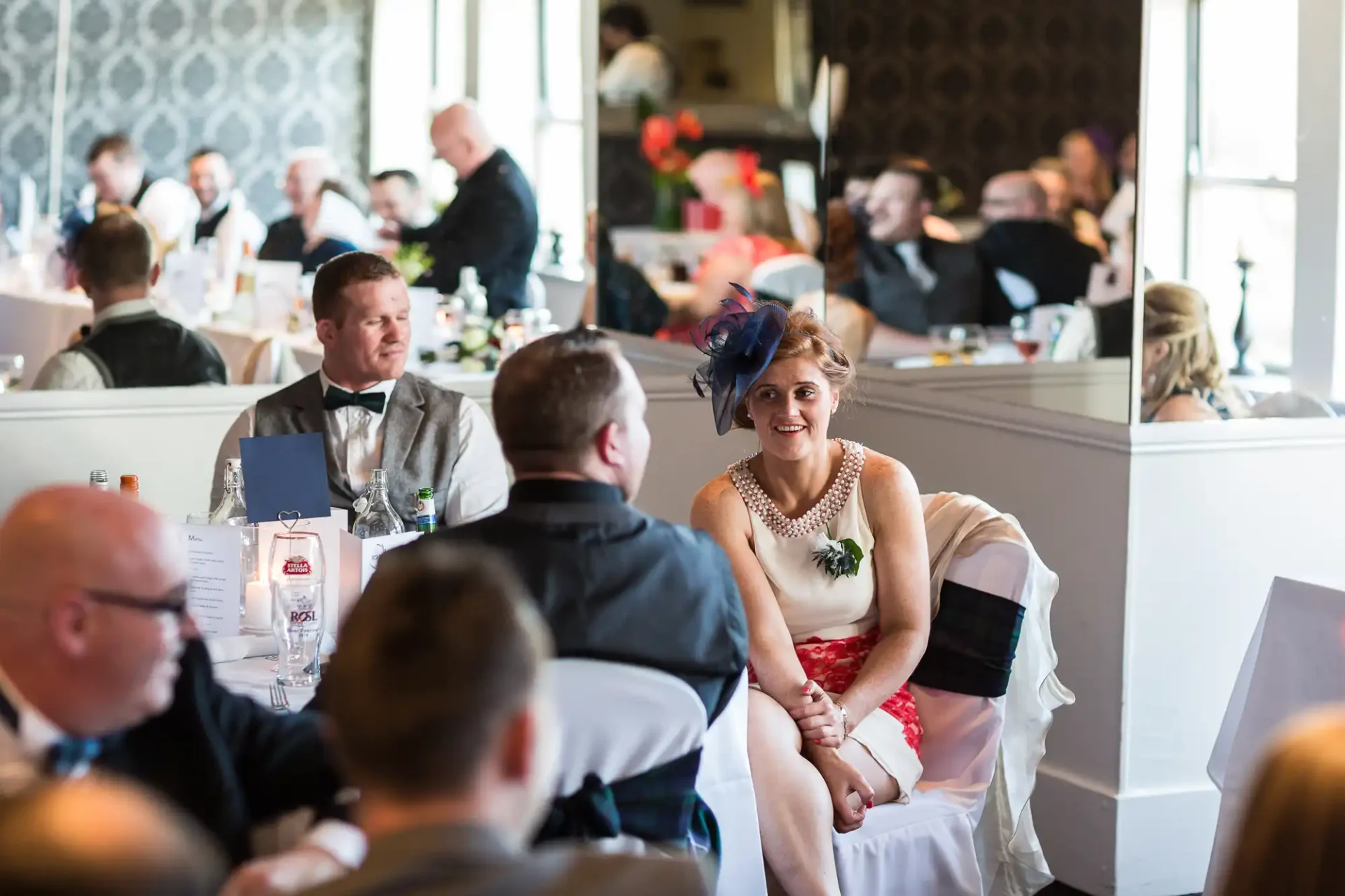 A bride in a white dress with a red sash sits at a wedding reception surrounded by guests in formal attire.
