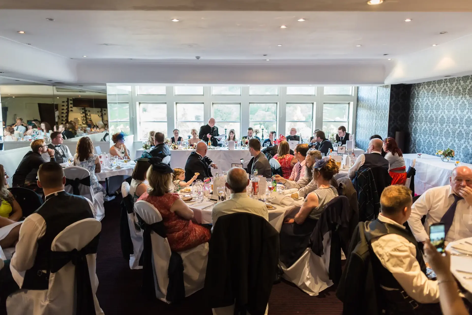 A wedding reception in a large room with guests seated at round tables, listening to a speaker at the front.