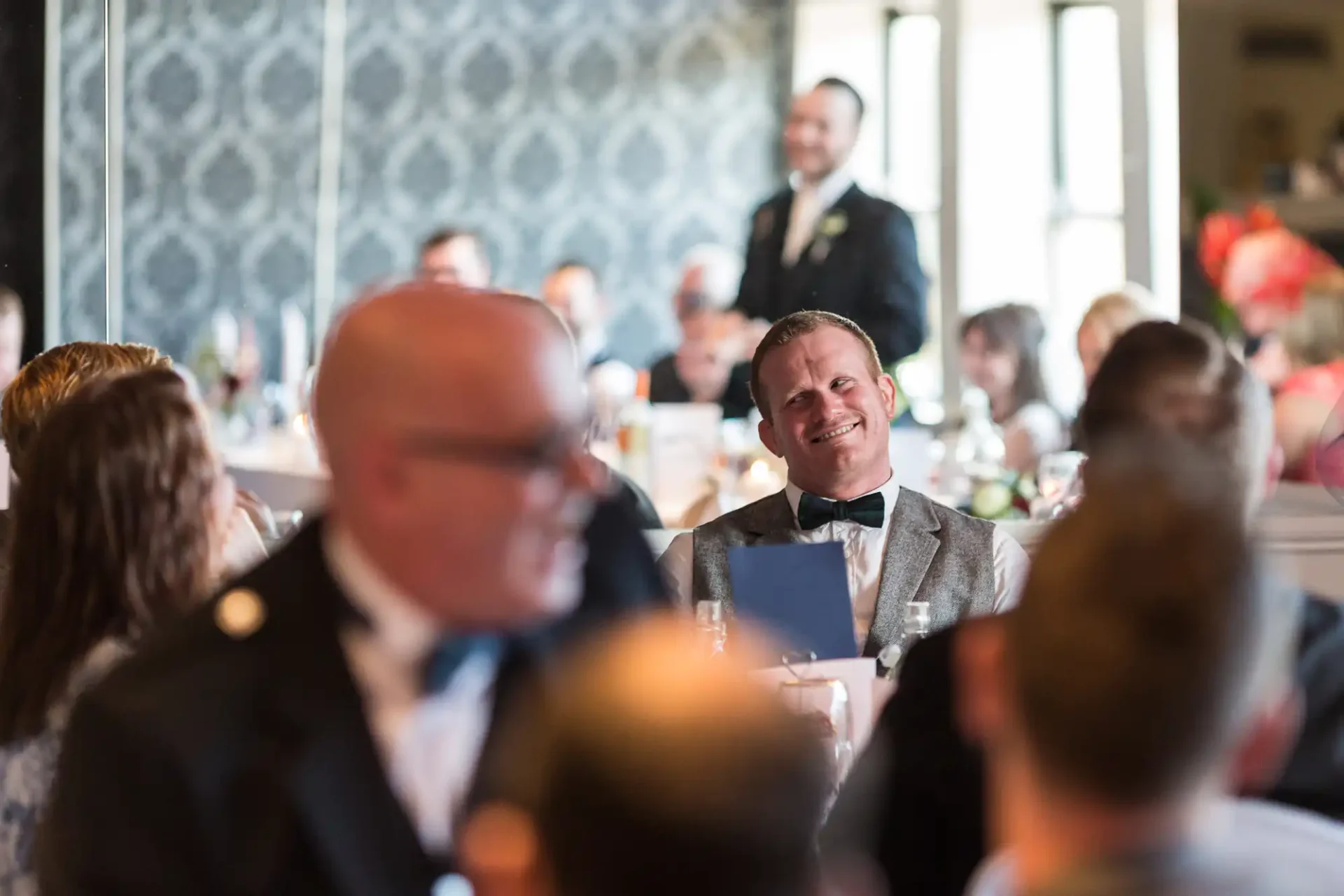 Man in a gray suit smiling at a wedding reception, surrounded by blurred guests in a room with elegant decor.