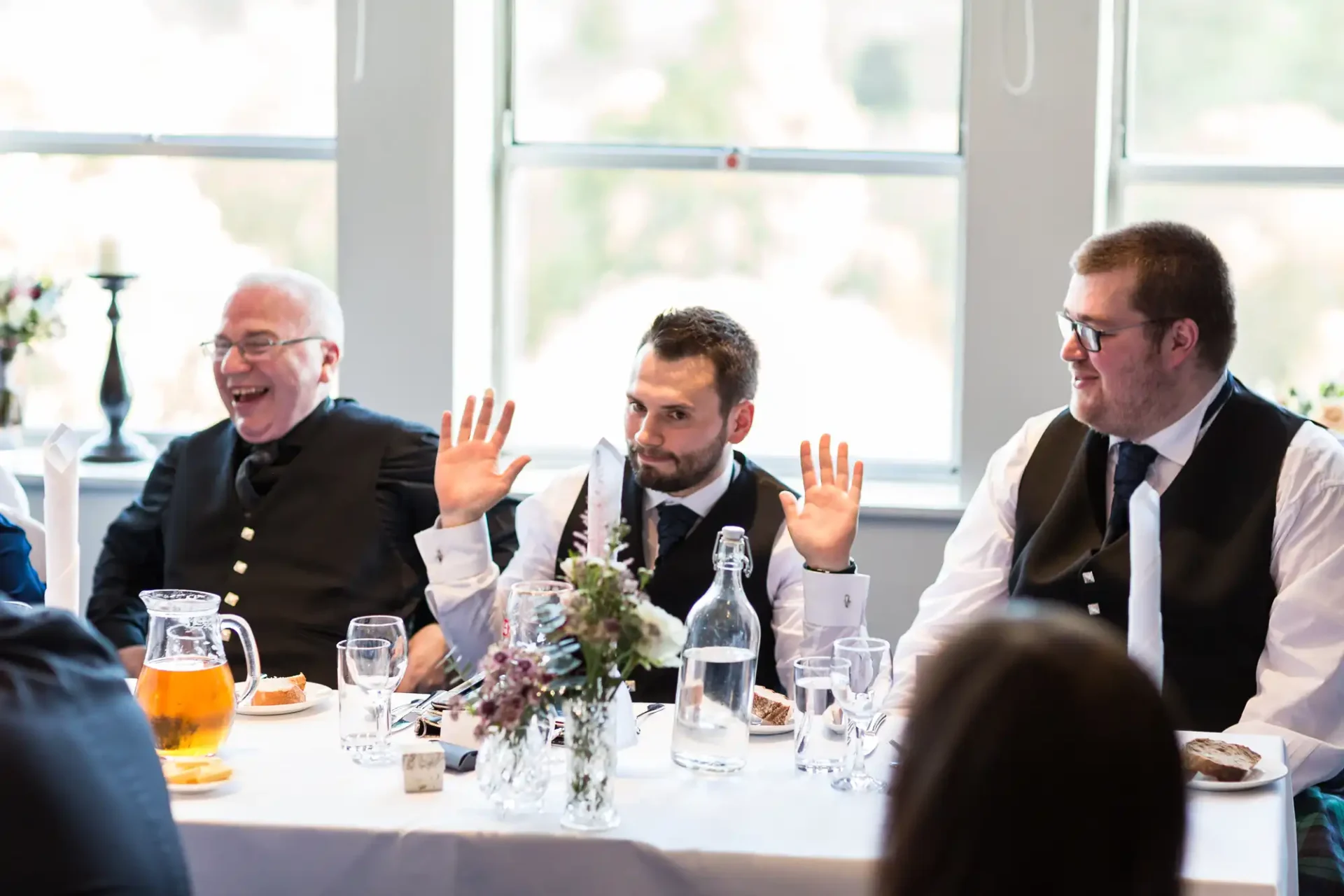Three men seated at a wedding reception table, laughing and gesturing with their hands, drinks and floral centerpiece on the table.