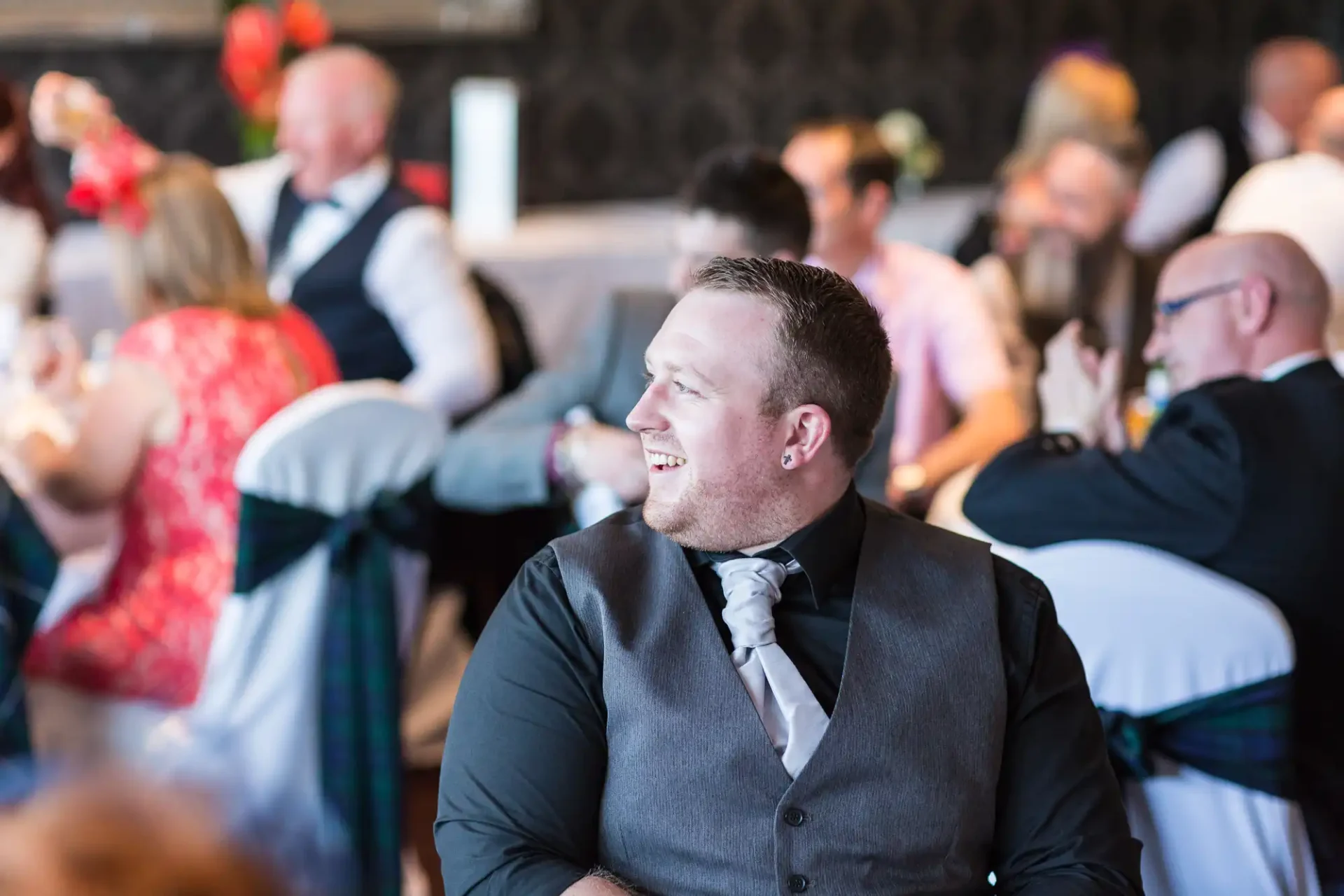 A man in a formal vest and tie smiling and looking to the side at a blurry, busy event with people in the background.