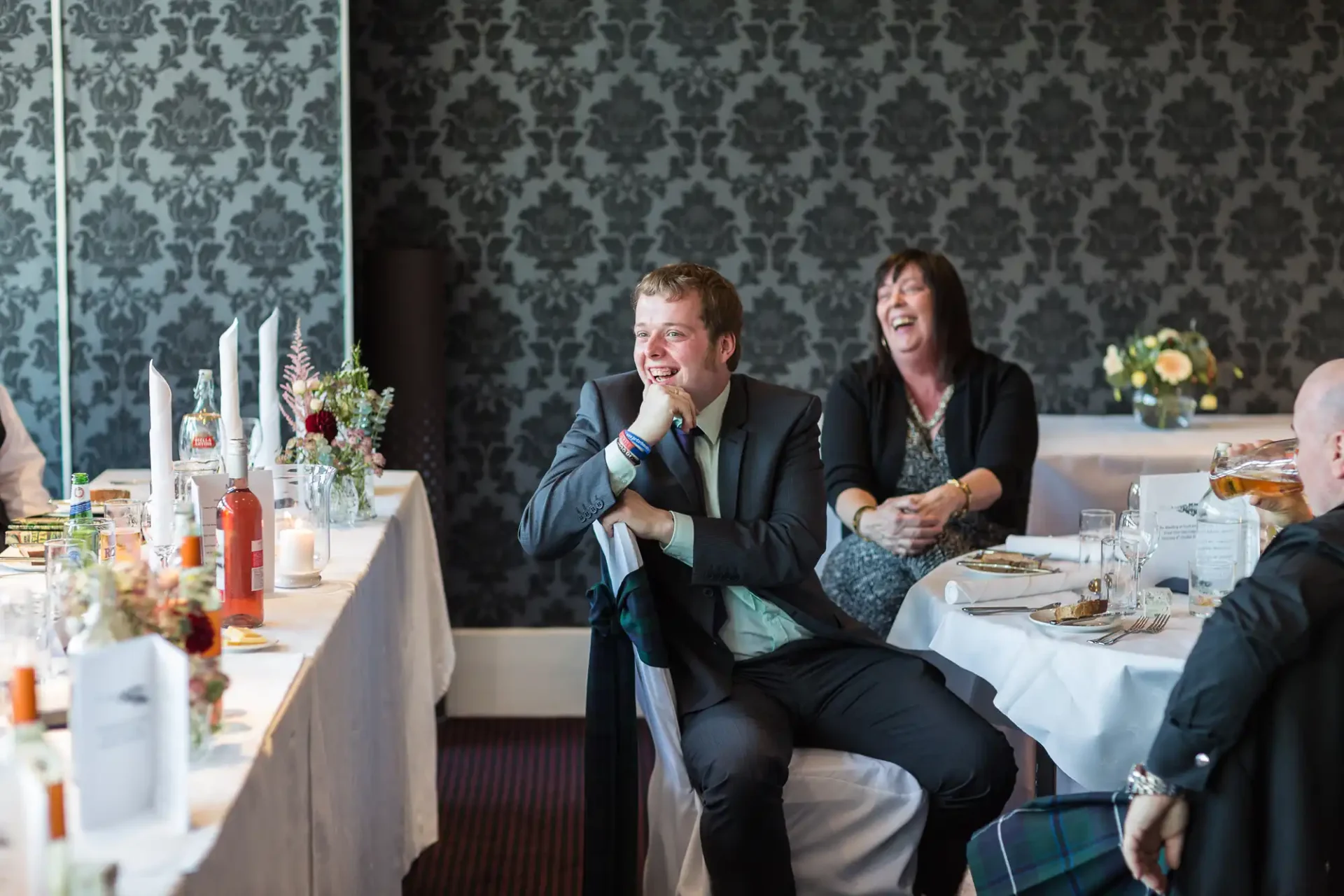 A man in a dark suit laughing at a wedding reception table, with a woman in black laughing beside him. decorative floral centerpieces and elegant table settings visible.