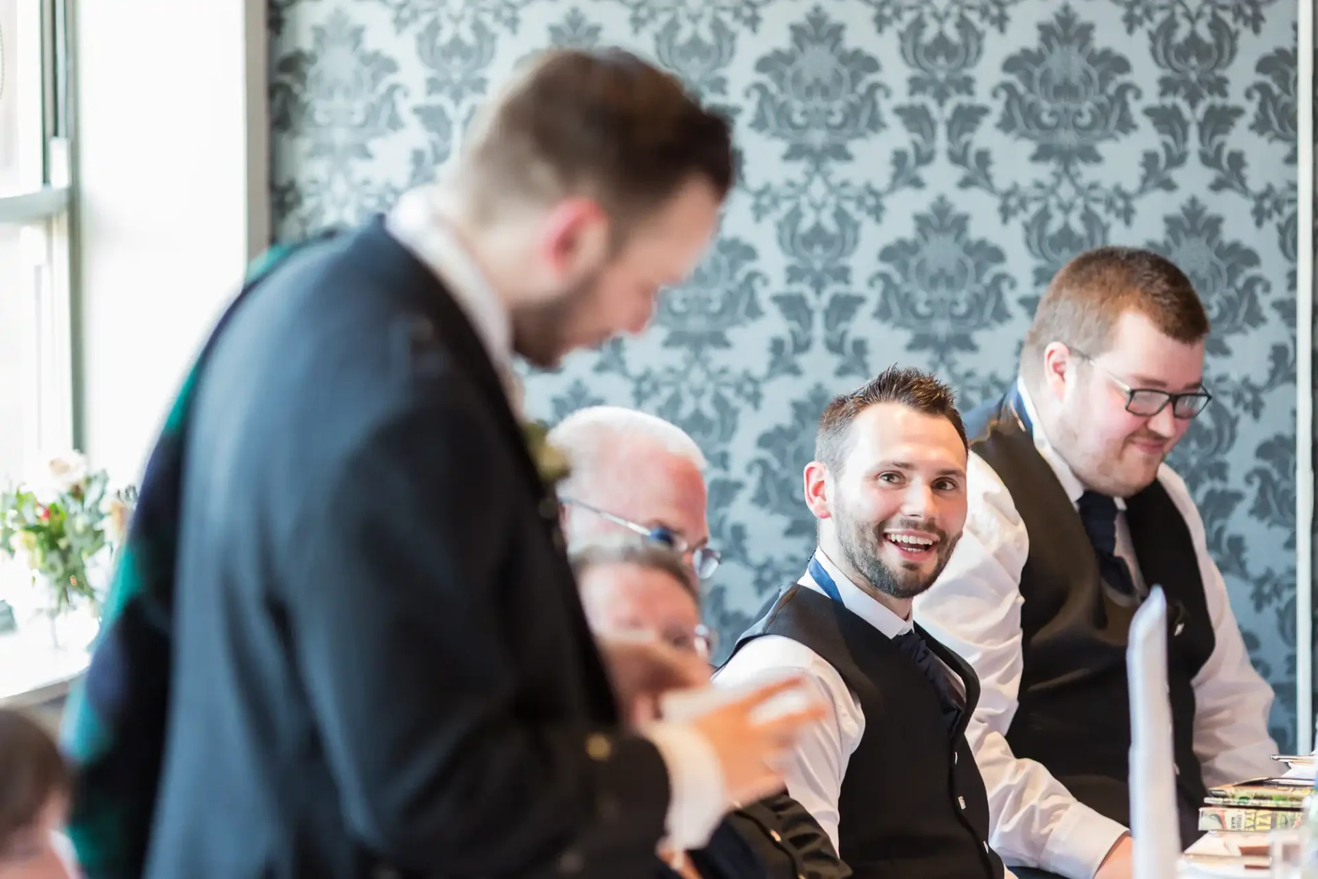 Group of men in formal wear smiling and interacting during an event in a room with elegant wallpaper and natural light.