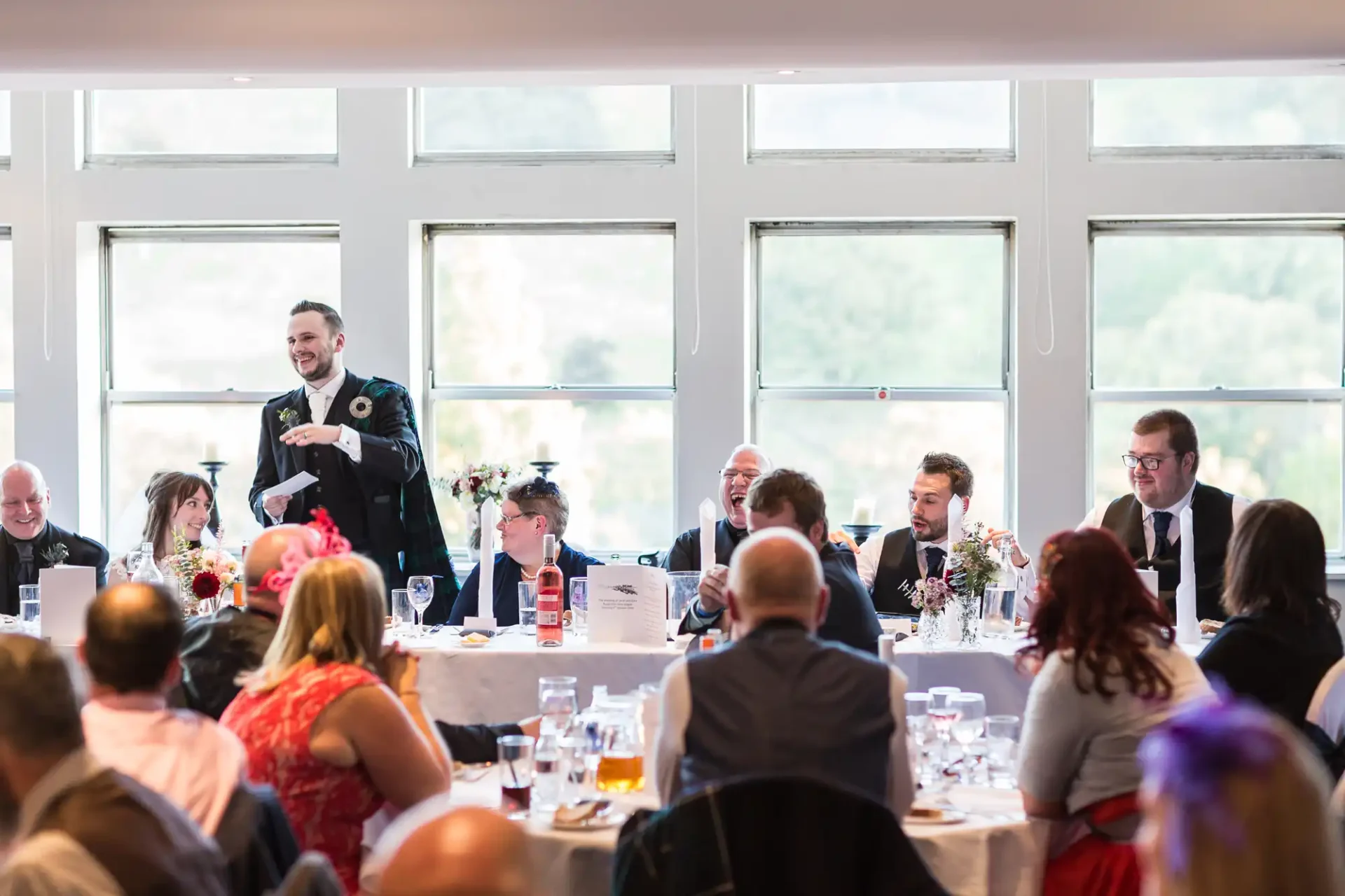 A man in a suit is giving a speech at a wedding reception, with guests seated at tables listening and smiling in a room with large windows.