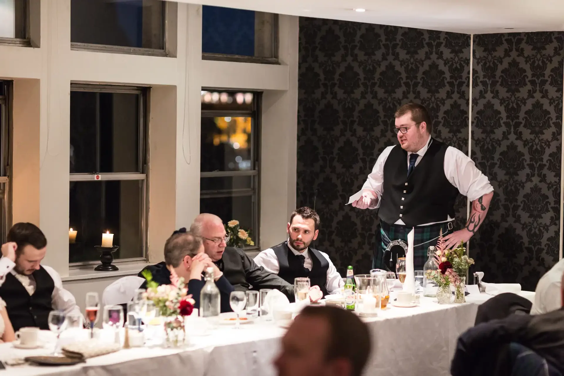 A waiter serving guests at a formal dinner party in an elegantly decorated room with large windows.
