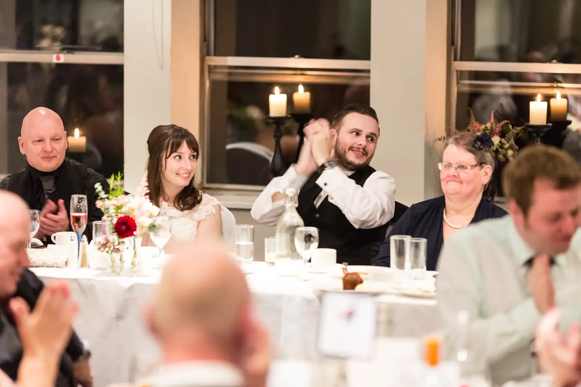 People at a wedding reception table, one man clapping and smiling, guests engaged in conversations.
