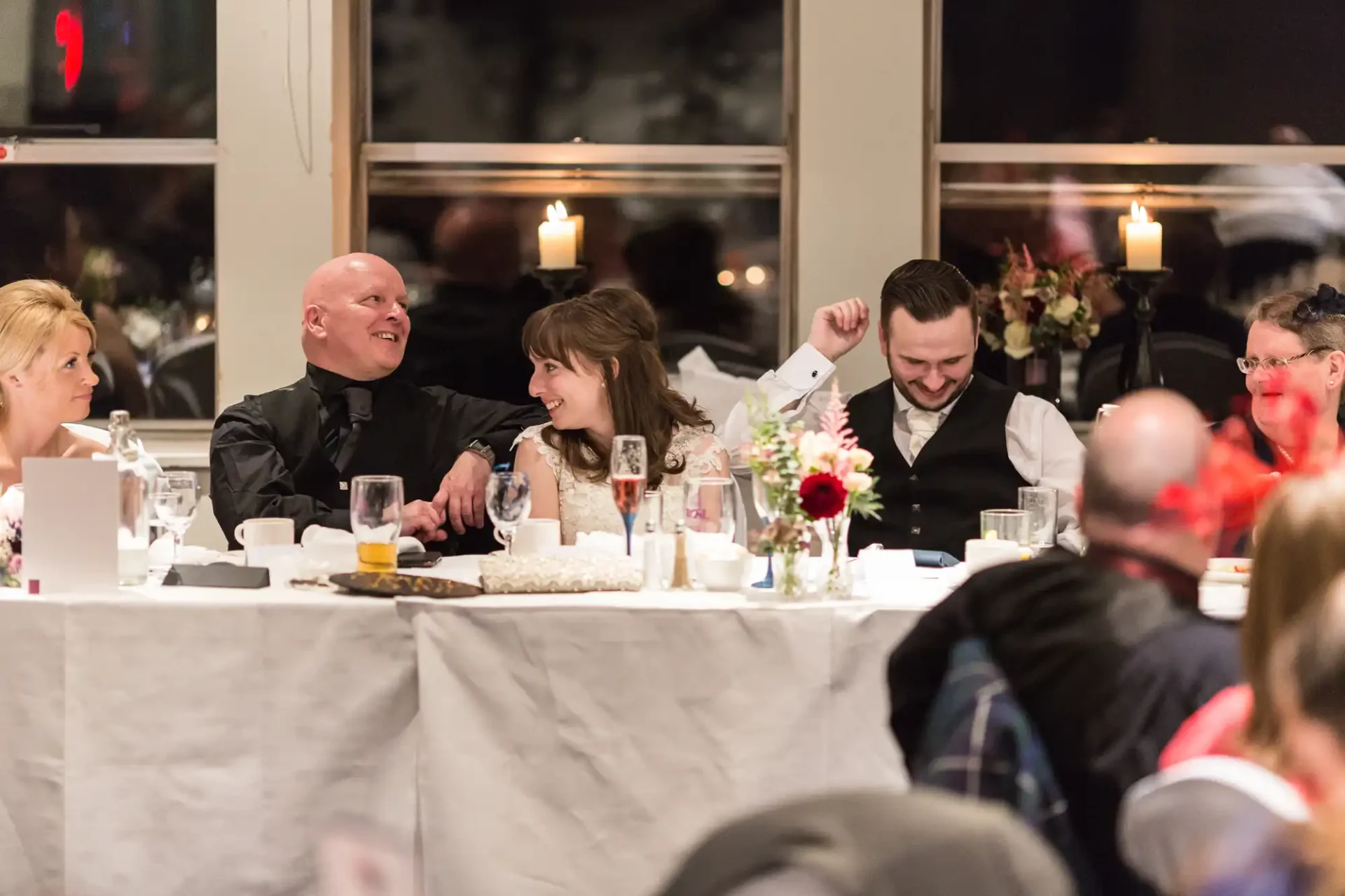 A joyful wedding reception scene showing a smiling bride and groom chatting with guests at a decorated table.