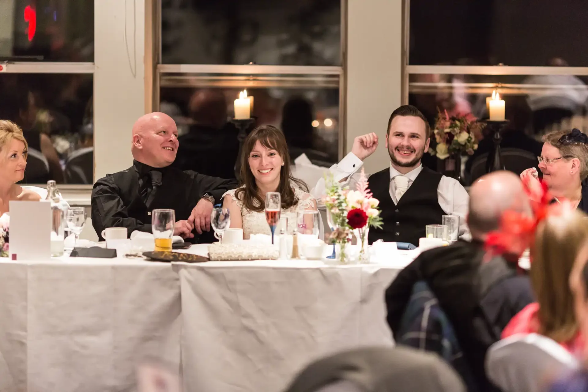 A joyful wedding reception scene with guests smiling and interacting at a decorated table. one man gestures excitedly.