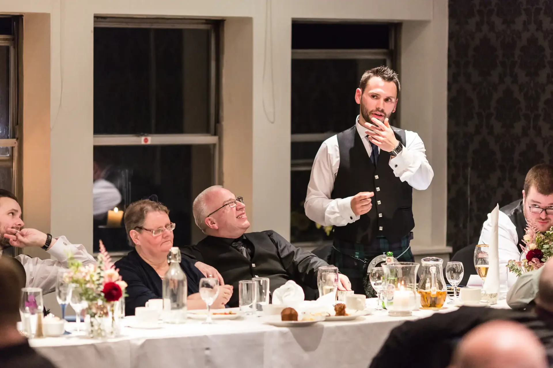 A waiter adjusting his tie while standing beside a table where a group of men sit, some laughing, during a dinner event in an elegant room.