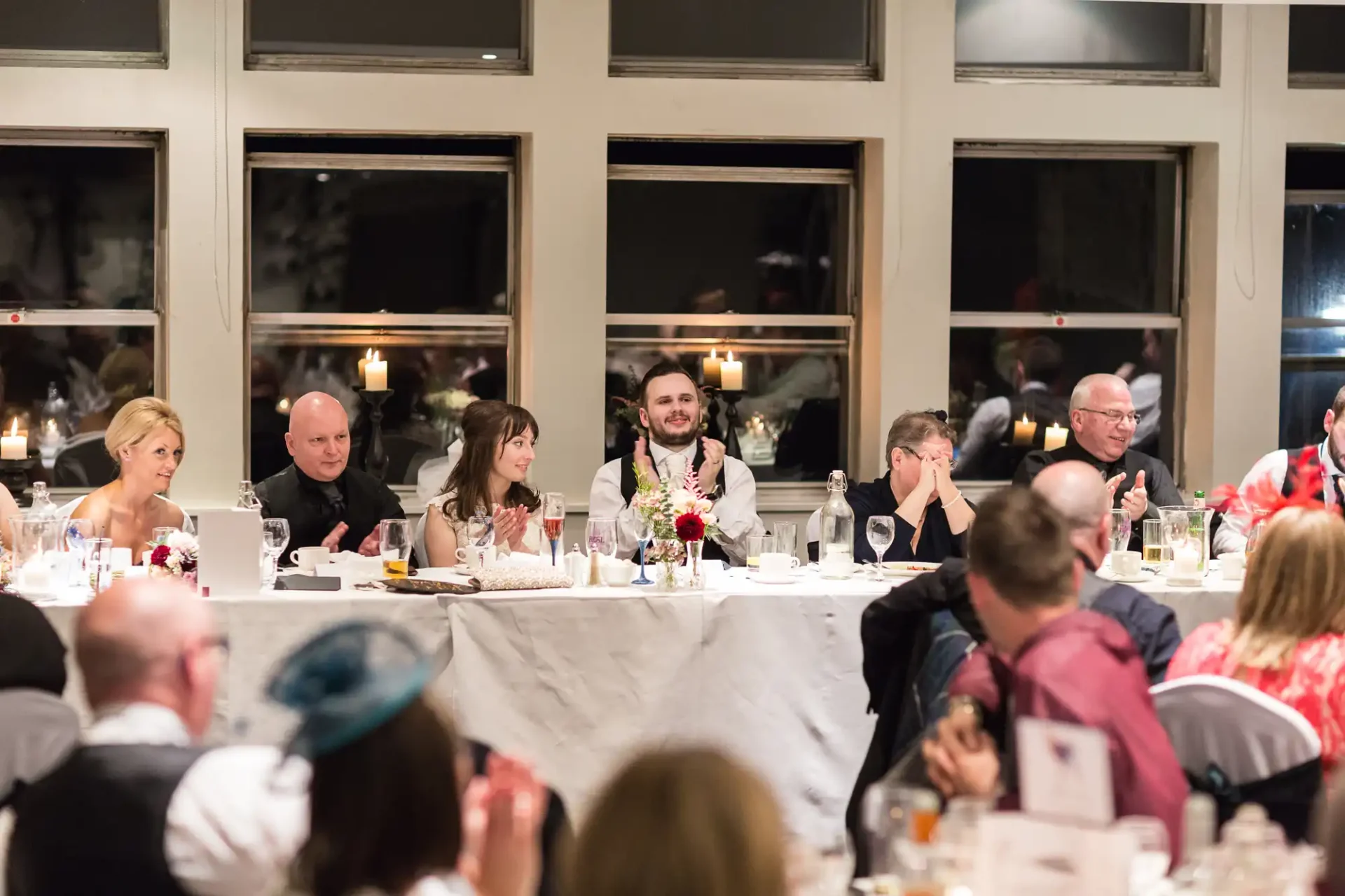 A formal dinner event with elegantly dressed guests seated at a long table, engaged in conversation, in a room with large windows.