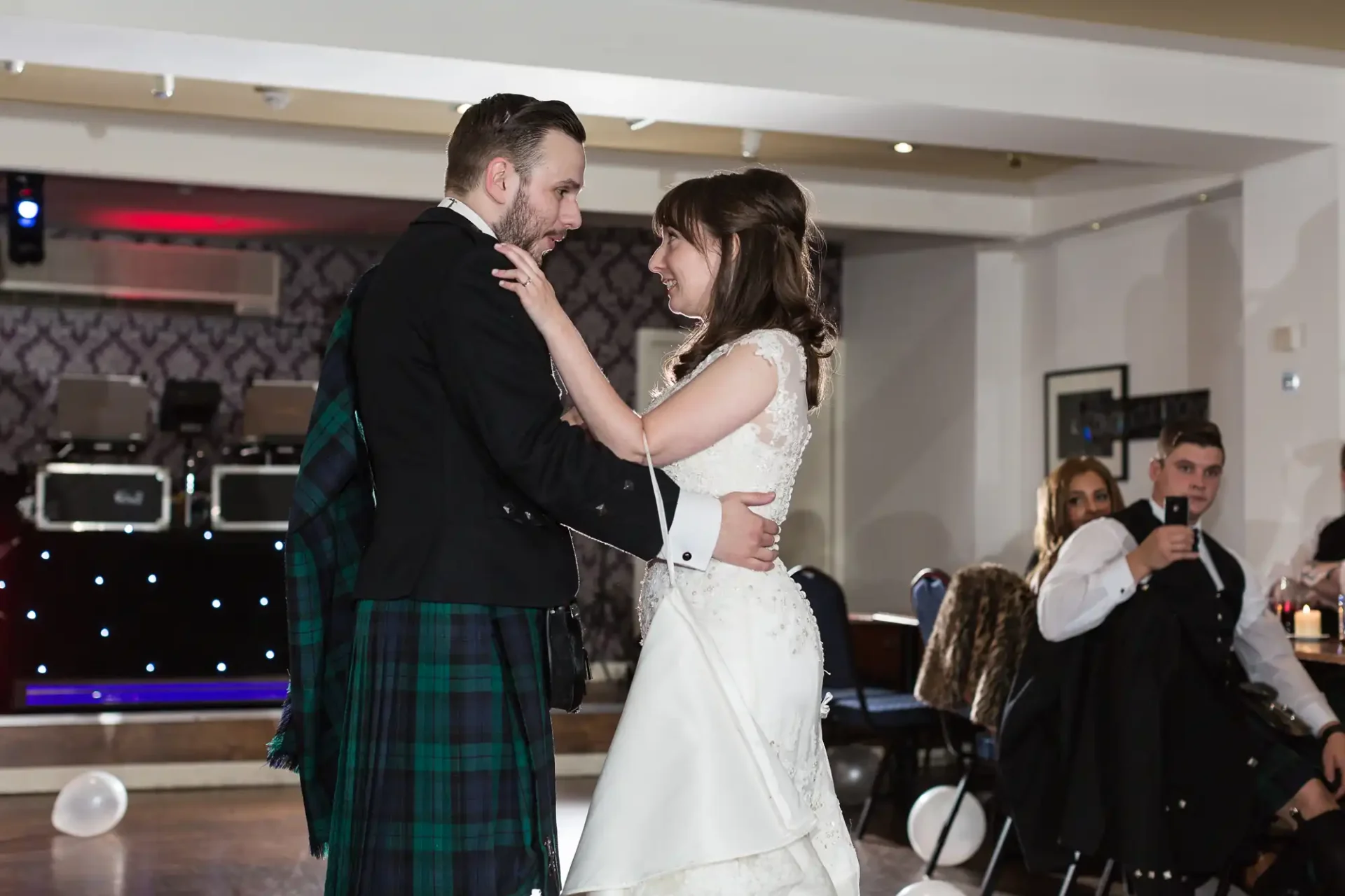 A bride and groom share a dance at their wedding reception, guests watching in the background.