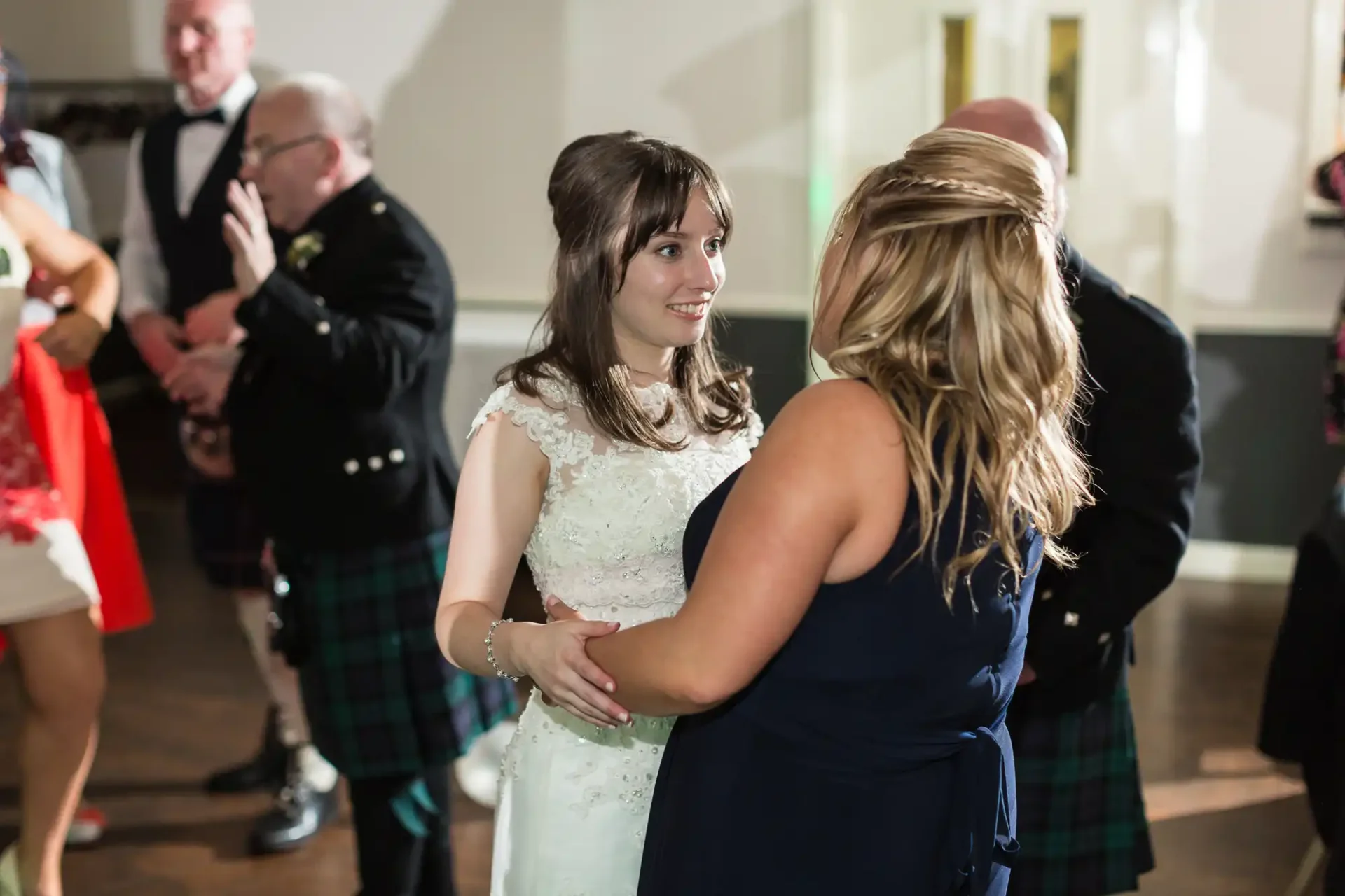 Two women in formal attire, one in a wedding dress, holding hands and smiling at each other at a dance floor with guests in background.