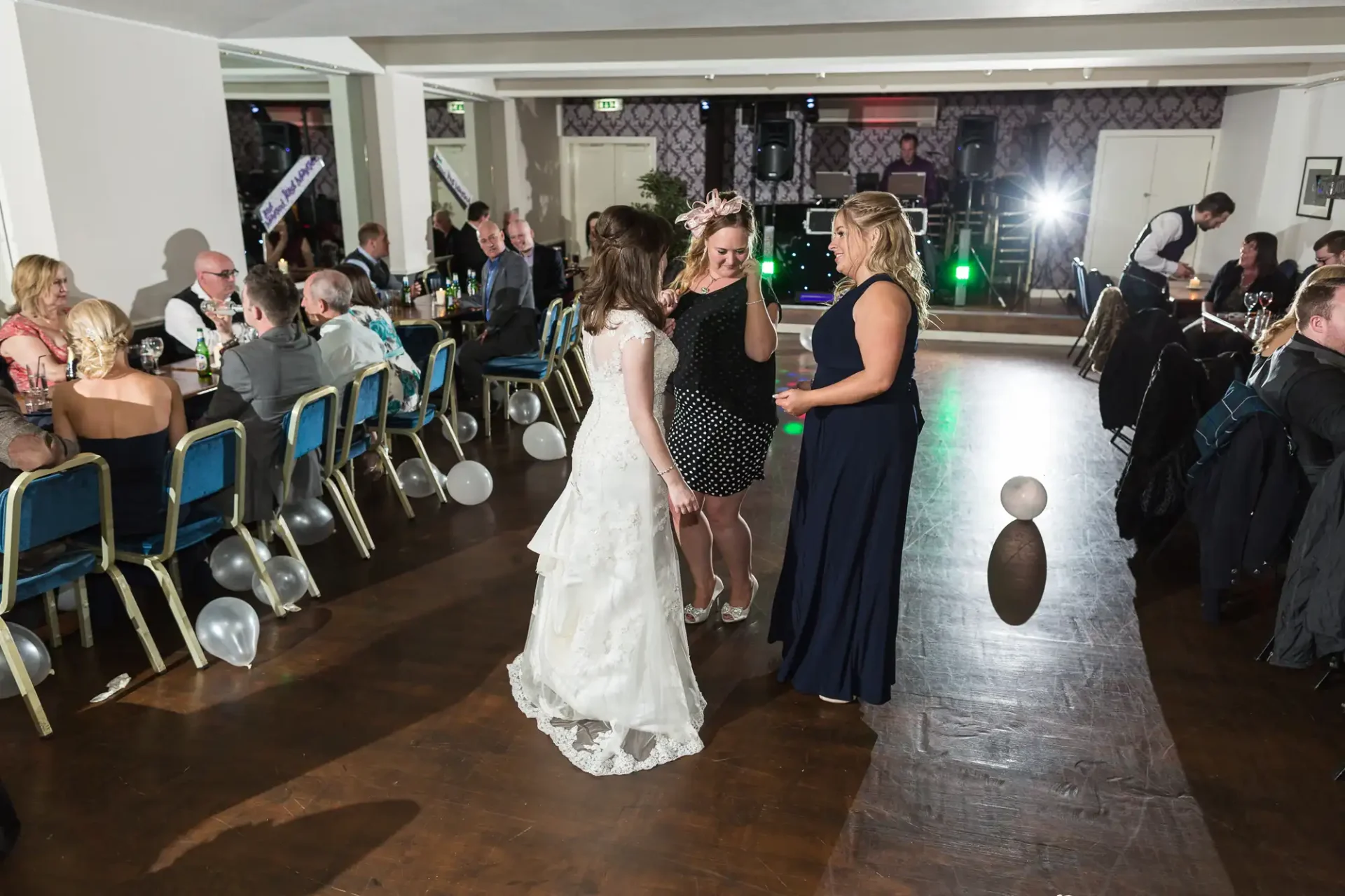 Three women, one in a white dress and two in evening gowns, chat on a dance floor at a festive event with seated guests around them.