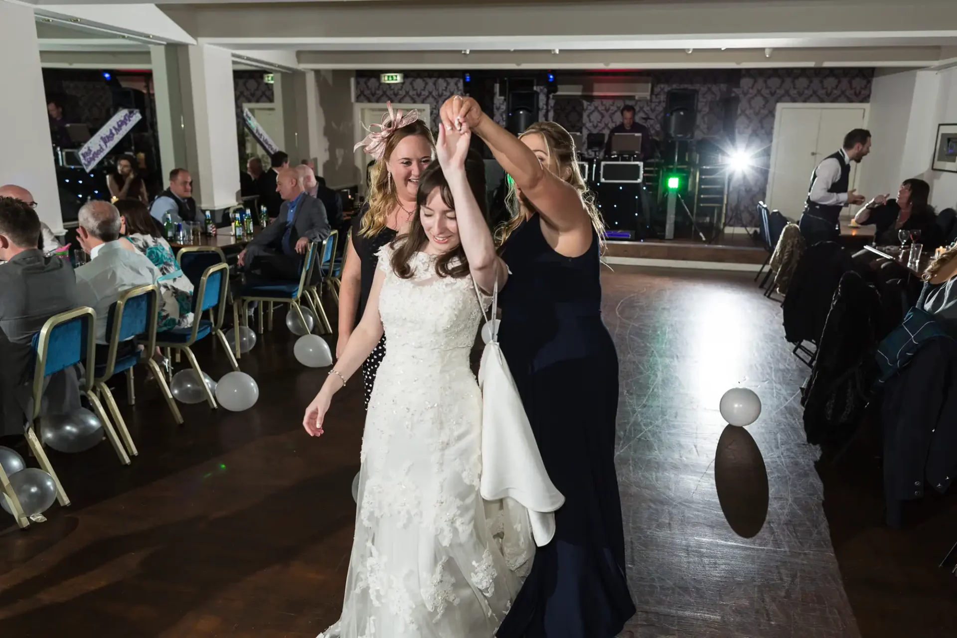 Two women in formal dresses dancing and smiling, holding hands raised in a celebratory gesture at a wedding reception with seated guests in the background.