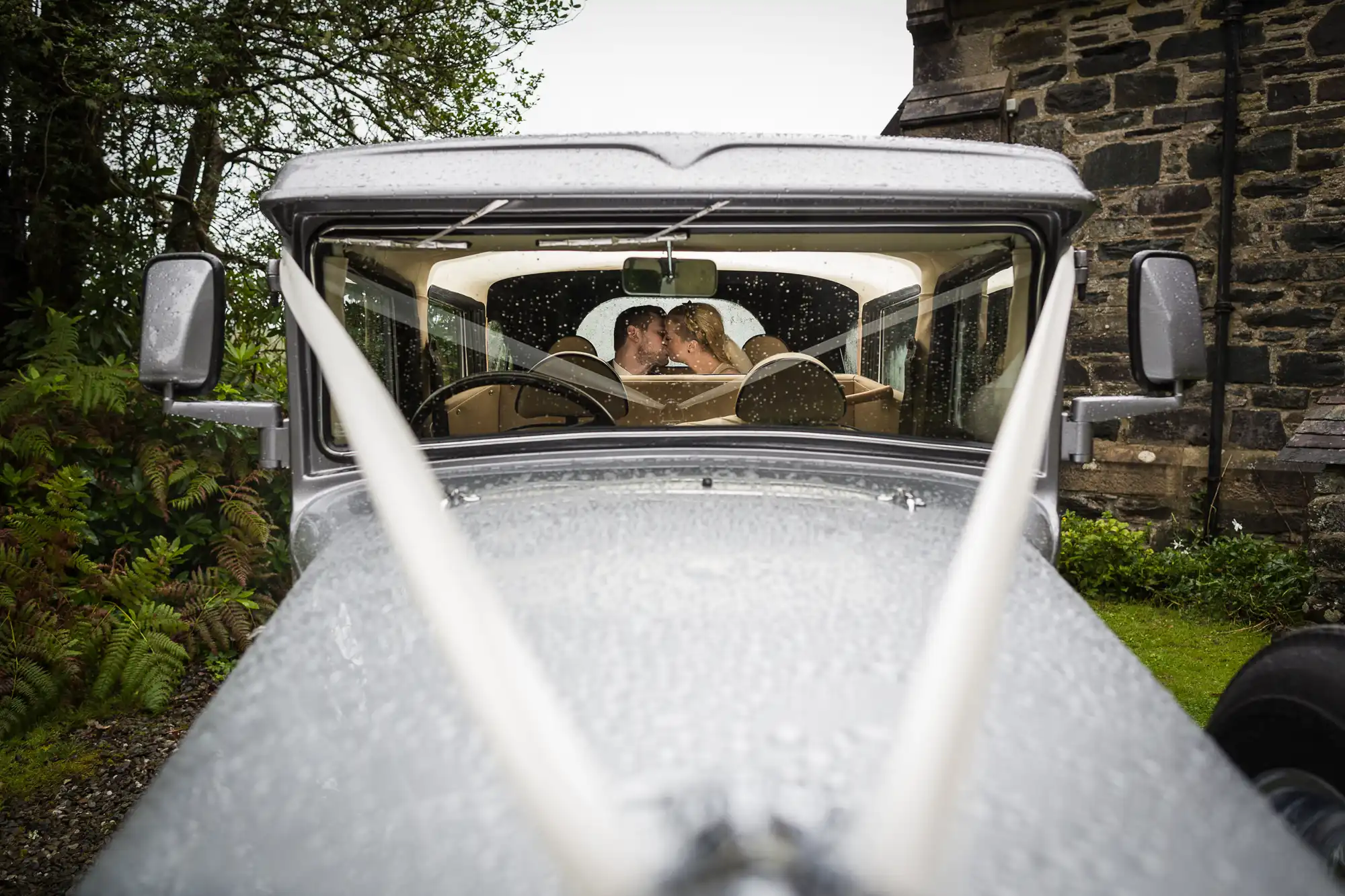 A couple kissing in the backseat of a vintage car on a rainy day, viewed from the vehicle's front by Aberfoyle Church photographer.
