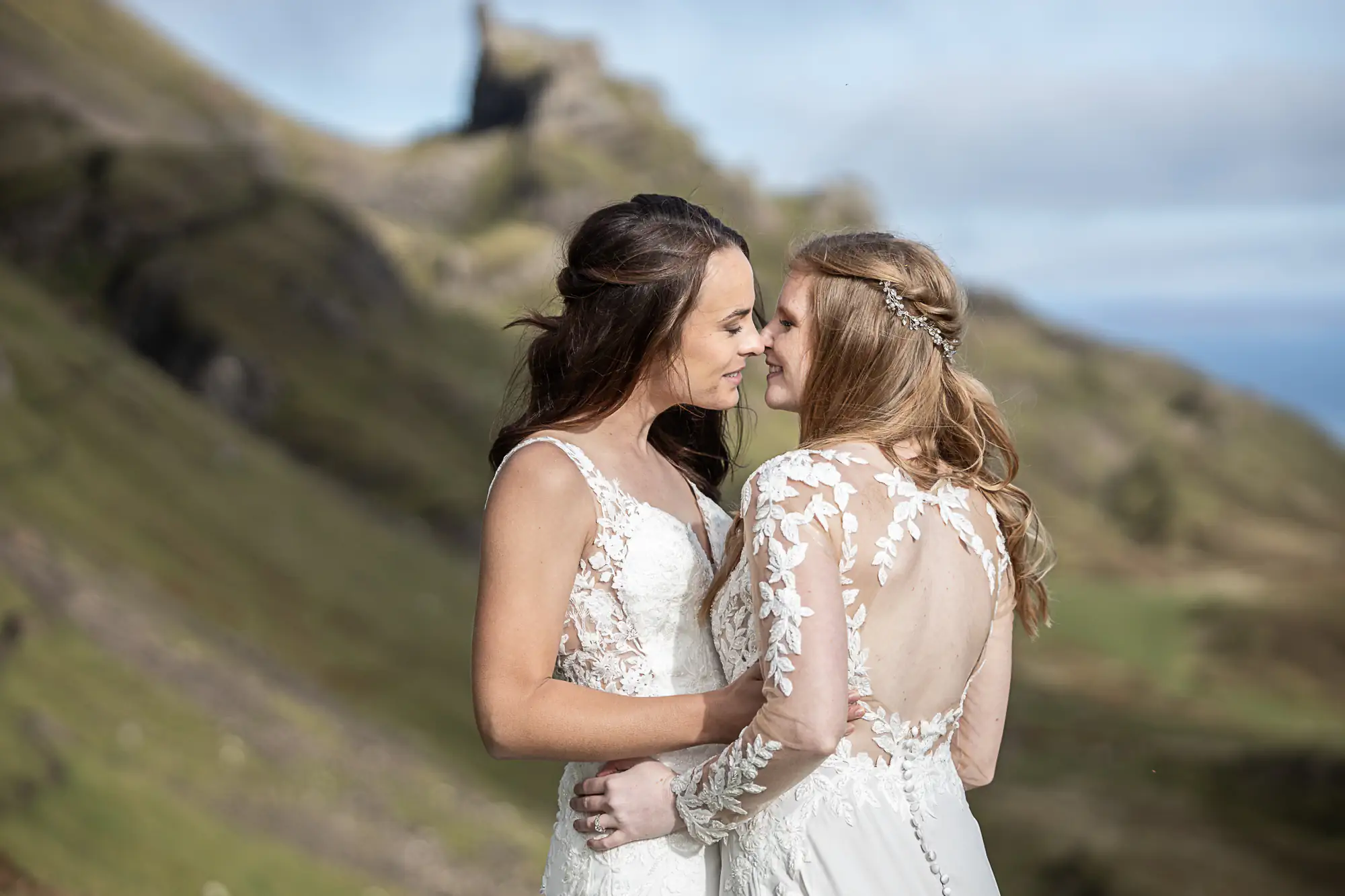 Two women in white dresses embrace and touch foreheads, standing outdoors with hilly landscape and cloudy sky in the background.