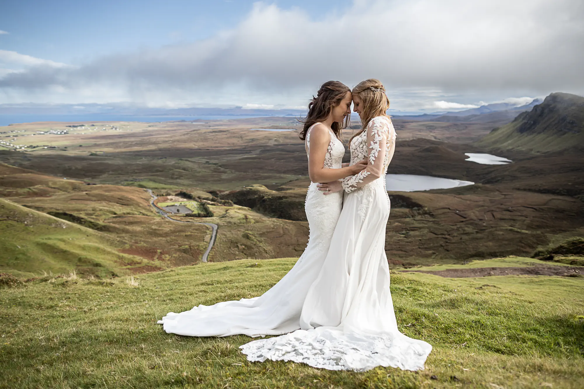 Two women in wedding dresses stand closely on a grassy hilltop with a scenic landscape of hills, a lake, and a cloudy sky in the background.