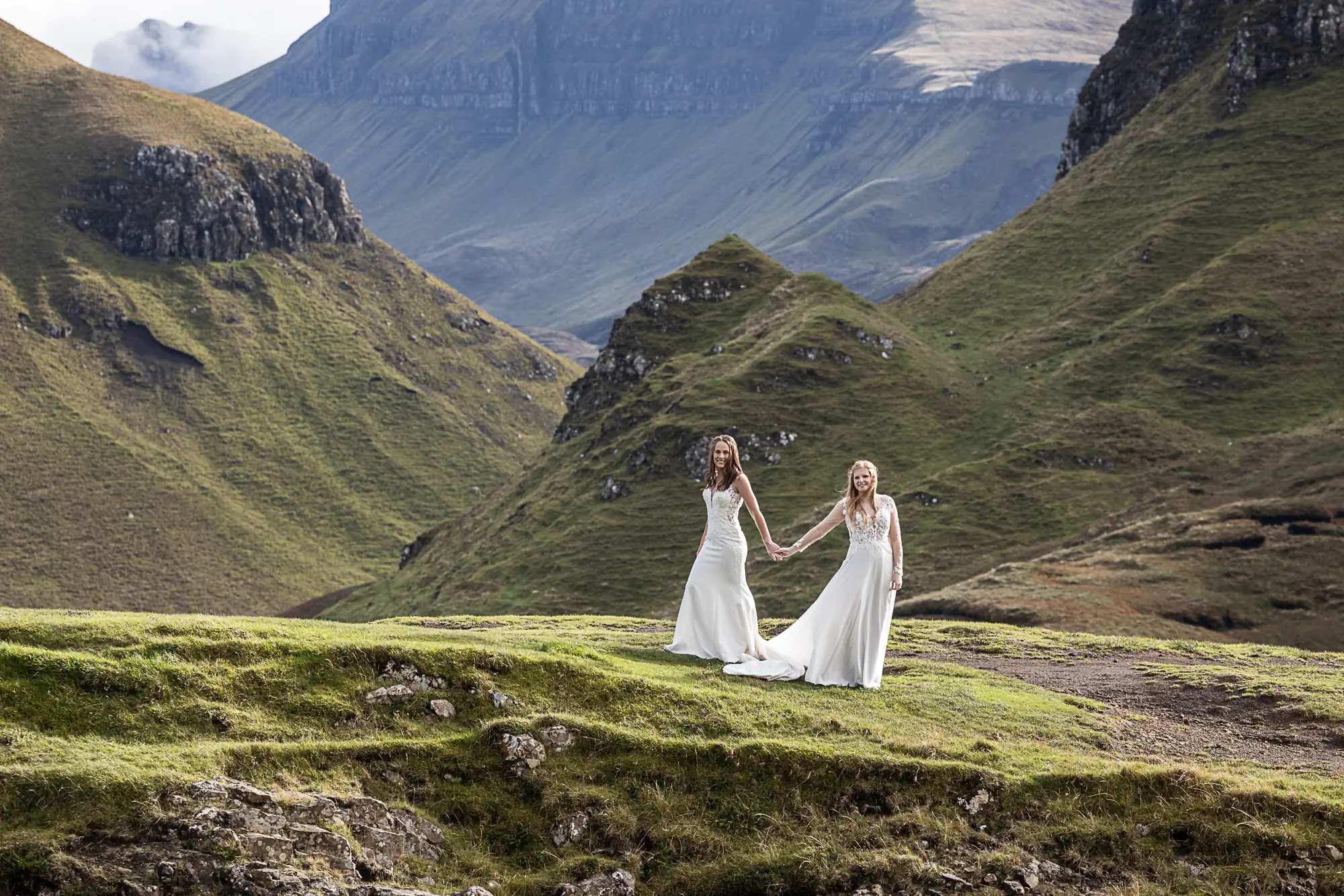 Two women in white dresses hold hands on a grassy hill with large mountains in the background on the Isle of Skye.