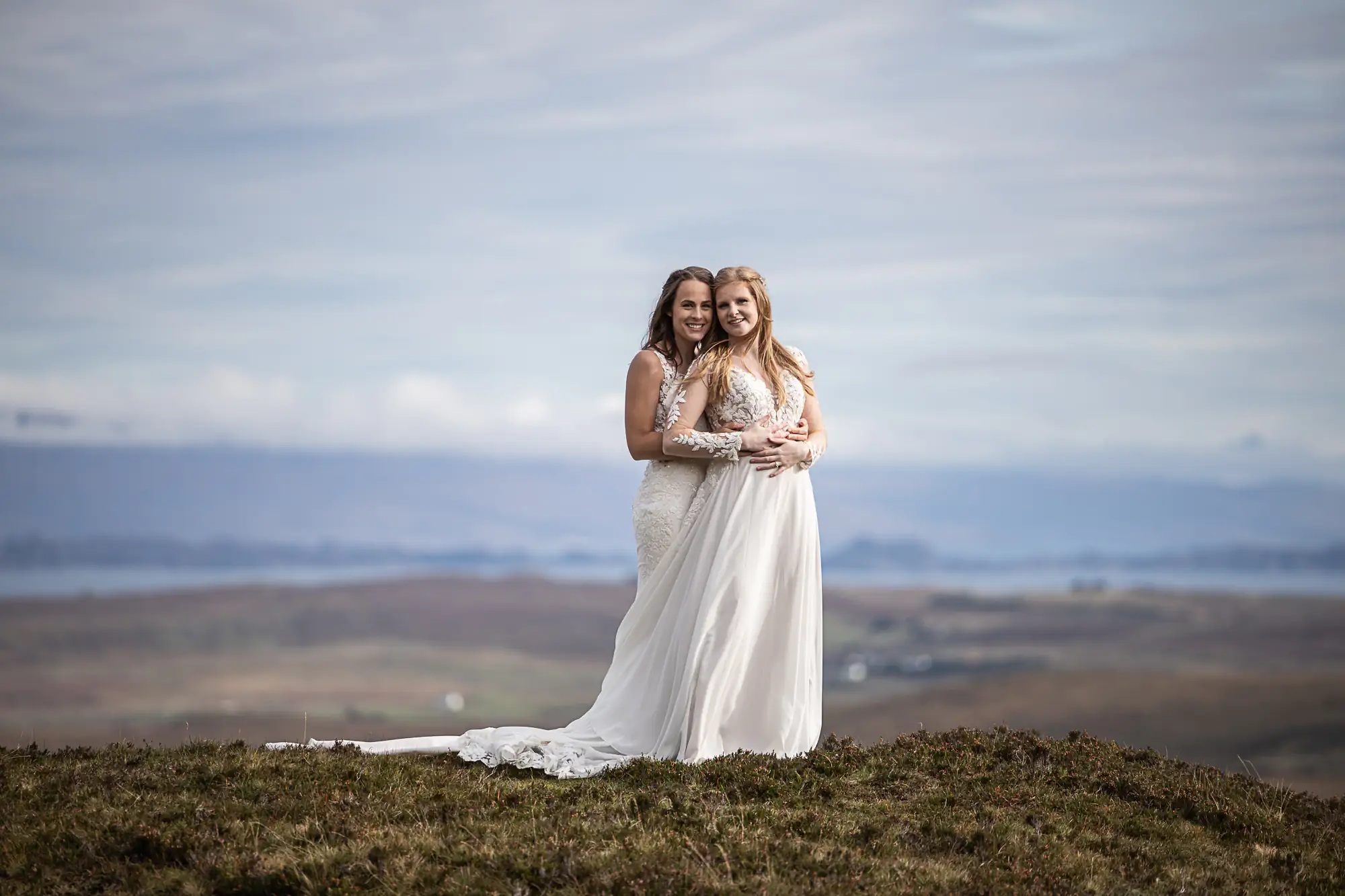 Two women in white wedding dresses stand close together on a grassy hilltop with a scenic, cloudy landscape in the background.