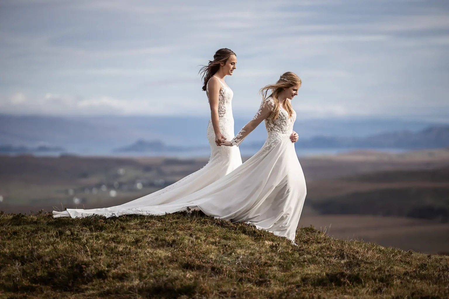 Scottish elopements photographer: Two brides in white wedding dresses hold hands while walking on a grassy hill with a scenic landscape in the background at The Quiraing, Isle of Skye.