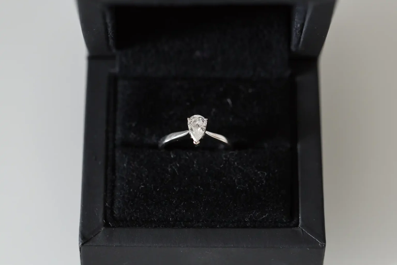 An engagement ring with a single diamond, set in a silver band, presented in an open black jewelry box.