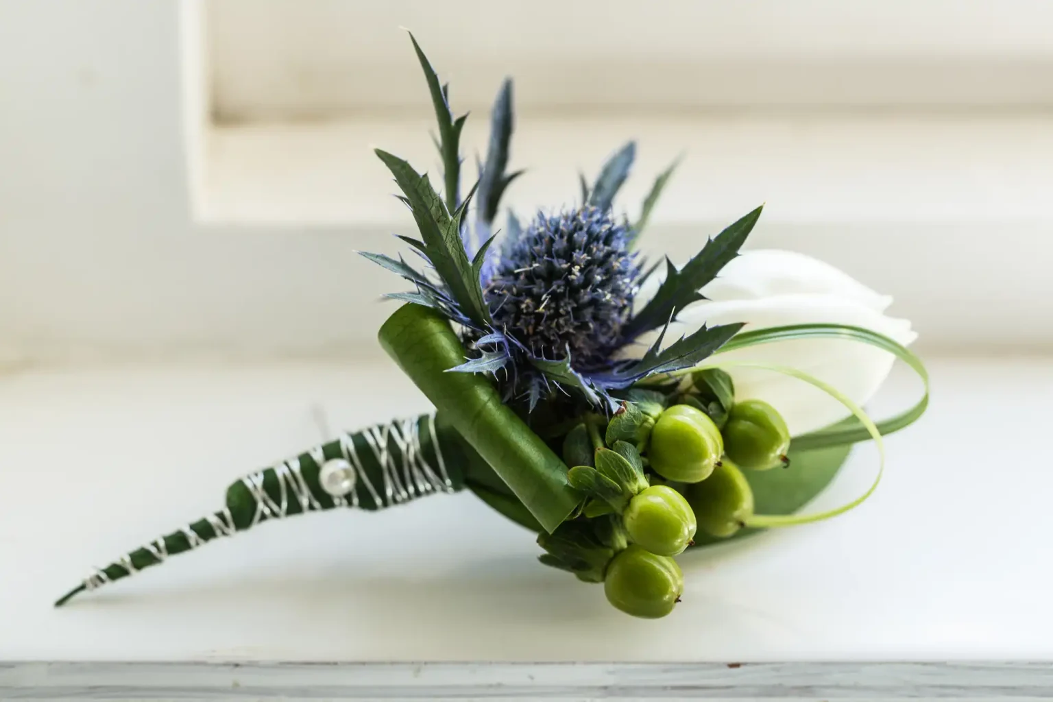A small, elegant bouquet featuring a blue thistle, green buds, and leafy stems wrapped with a white ribbon on a white surface.