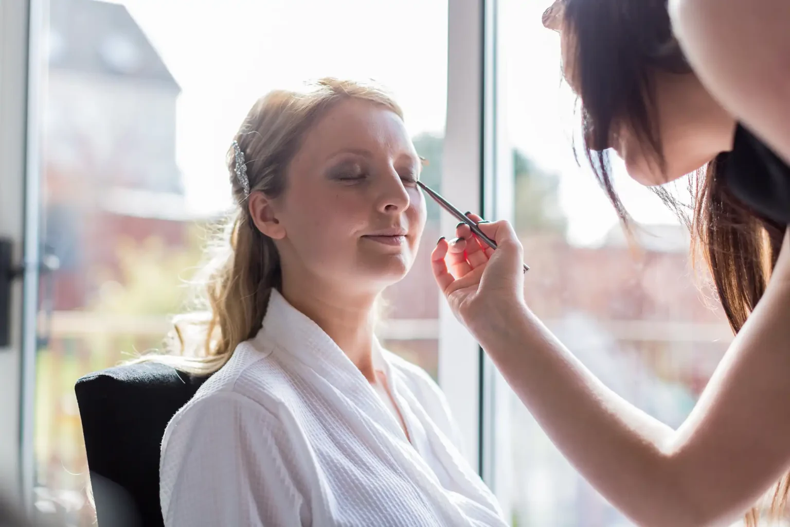 A woman having makeup applied by another person in a sunlit room by a large window.