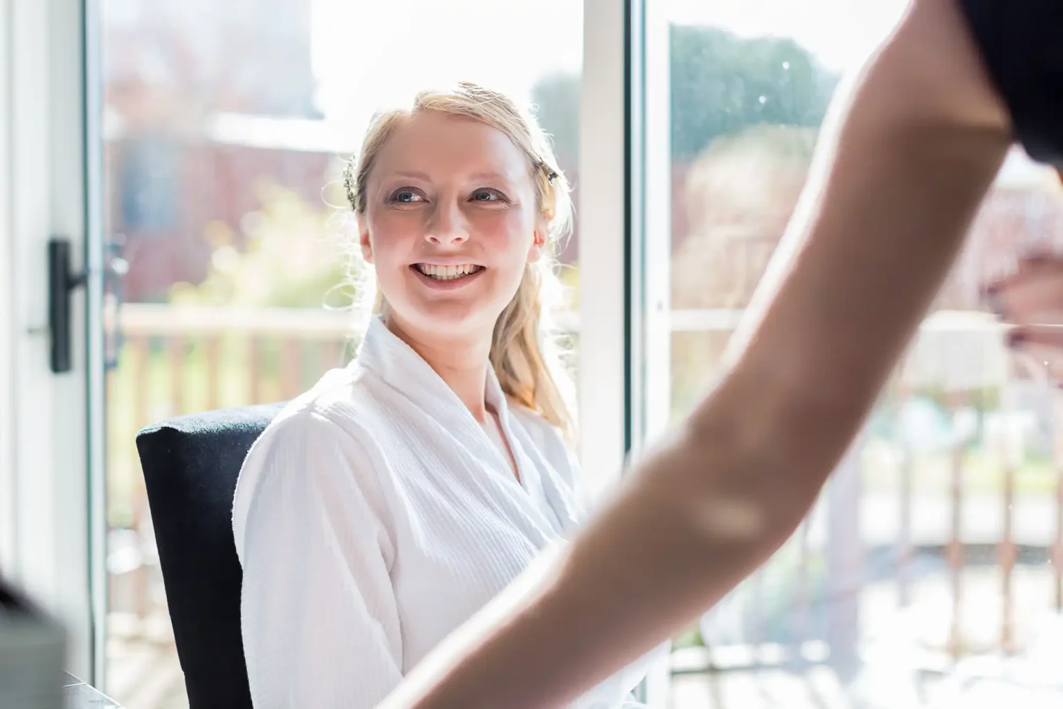 A smiling woman in a white robe looking at someone indoors with bright daylight coming from windows in the background.