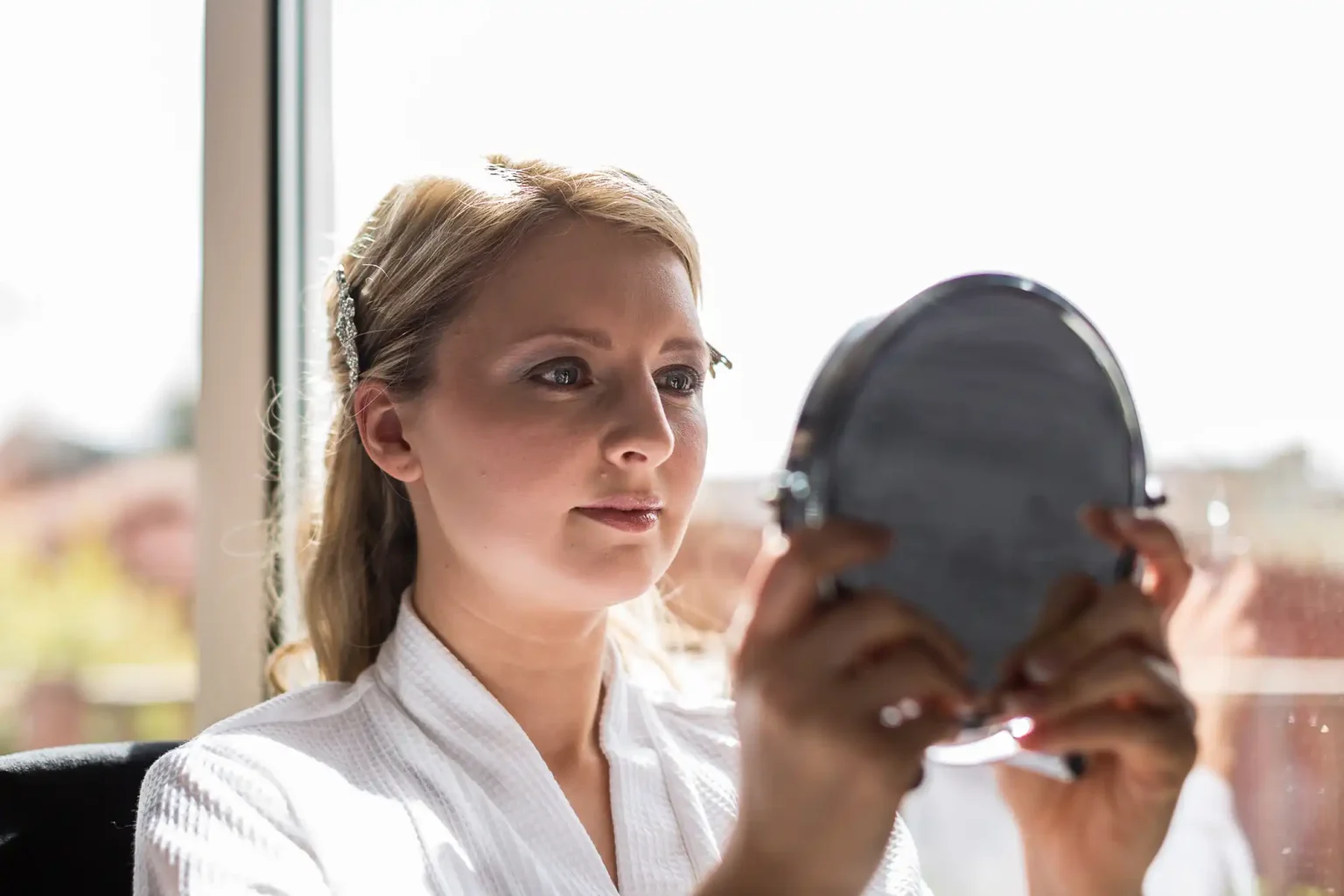 A woman in a white robe holding a handheld mirror, looking at her reflection with a focused expression, in a room with bright natural light.