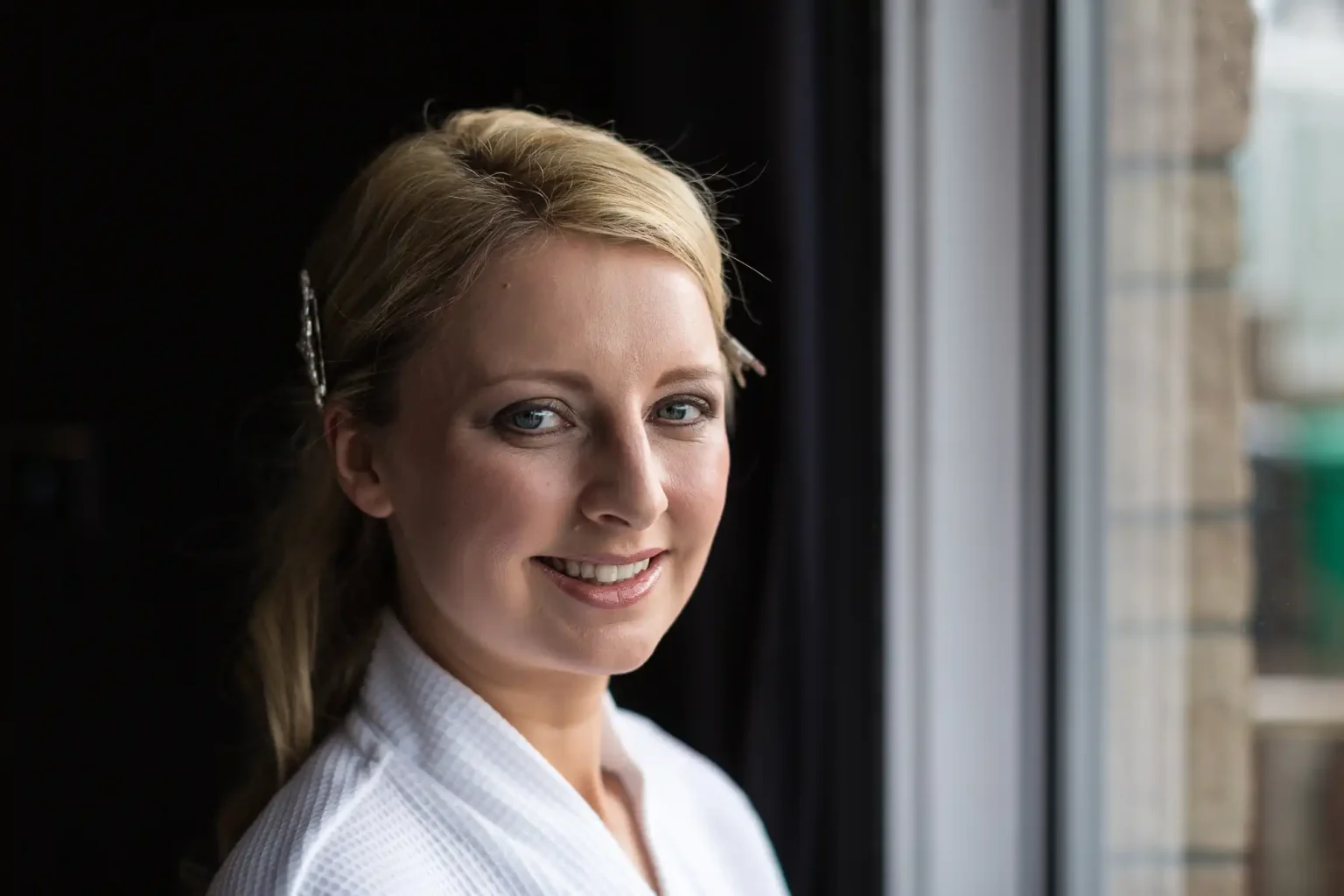 Portrait of a smiling caucasian woman with blonde hair, wearing a white robe, standing by a window.