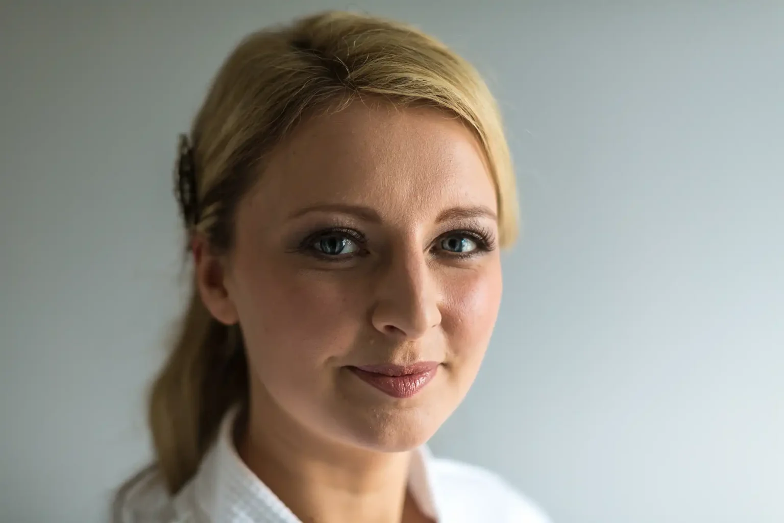 Close-up portrait of a smiling woman with blond hair, wearing a white shirt against a light gray background.