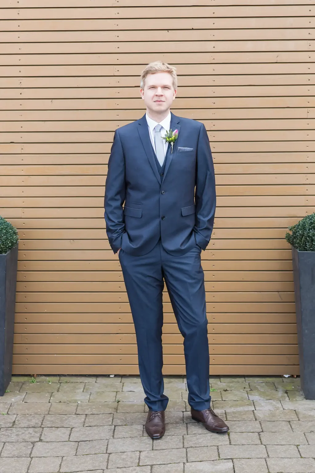 A man in a dark blue suit and tie with a boutonniere, standing upright against a wooden slat background.